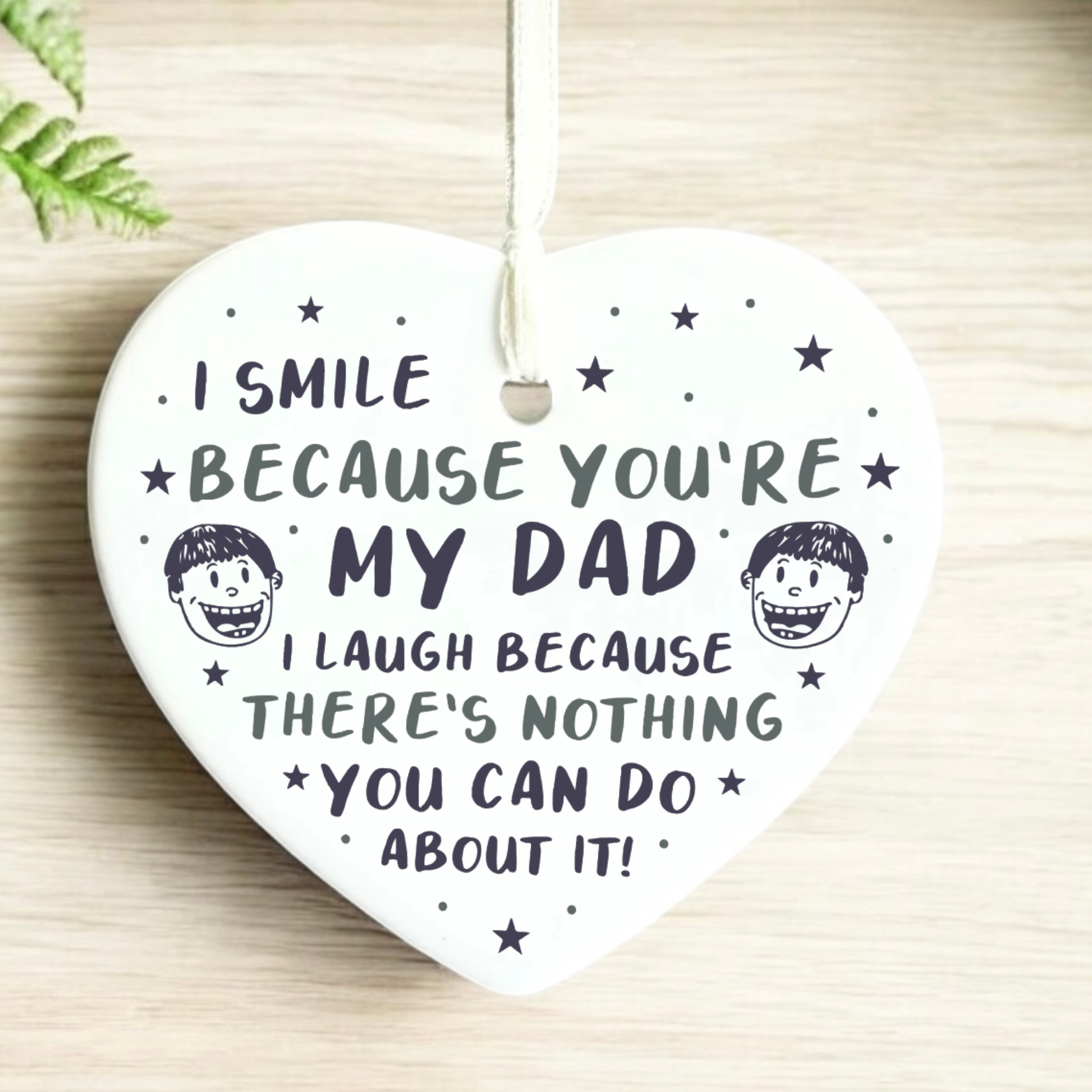 A close-up shot of a white ceramic heart-shaped hanging ornament held in two hands. The ornament displays a funny message that reads, "I smile because you're my dad. I laugh because there's nothing you can do about it!", decorated with stars and small illustrated cartoon faces.