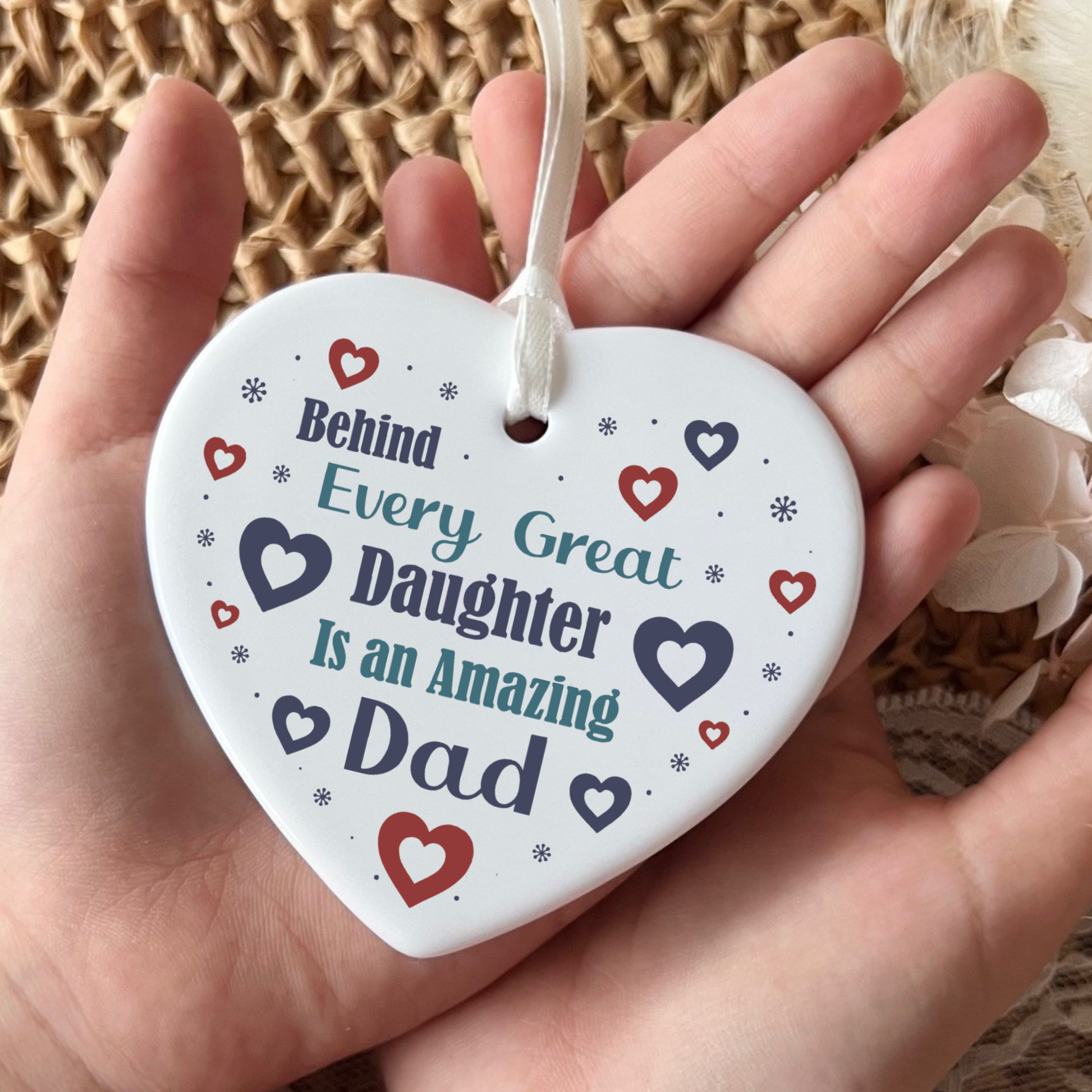 A close-up shot of a white ceramic heart-shaped hanging ornament displaying the text "Behind Every Great Daughter Is an Amazing Dad" surrounded by small red and navy hearts and tiny star patterns, complete with a white hanging ribbon.