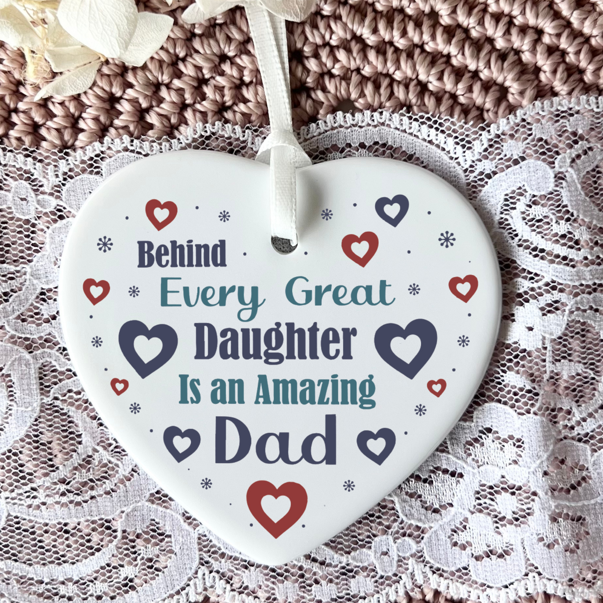 A close-up shot of a white ceramic heart-shaped hanging ornament displaying the text "Behind Every Great Daughter Is an Amazing Dad" surrounded by small red and navy hearts and tiny star patterns, complete with a white hanging ribbon.