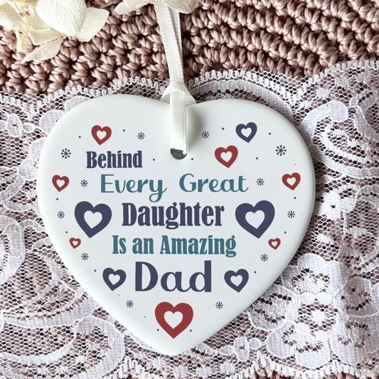 A close-up shot of a white ceramic heart-shaped hanging ornament displaying the text "Behind Every Great Daughter Is an Amazing Dad" surrounded by small red and navy hearts and tiny star patterns, complete with a white hanging ribbon.