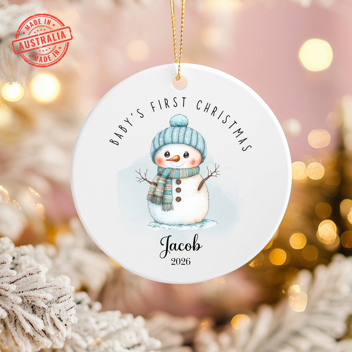 A personalized round white Christmas ornament featuring a watercolor snowman wearing a blue knit hat and scarf. The ornament is printed with the text 'BABY'S FIRST CHRISTMAS' at the top and 'Jacob 2026' at the bottom, complete with a gold hanging loop.