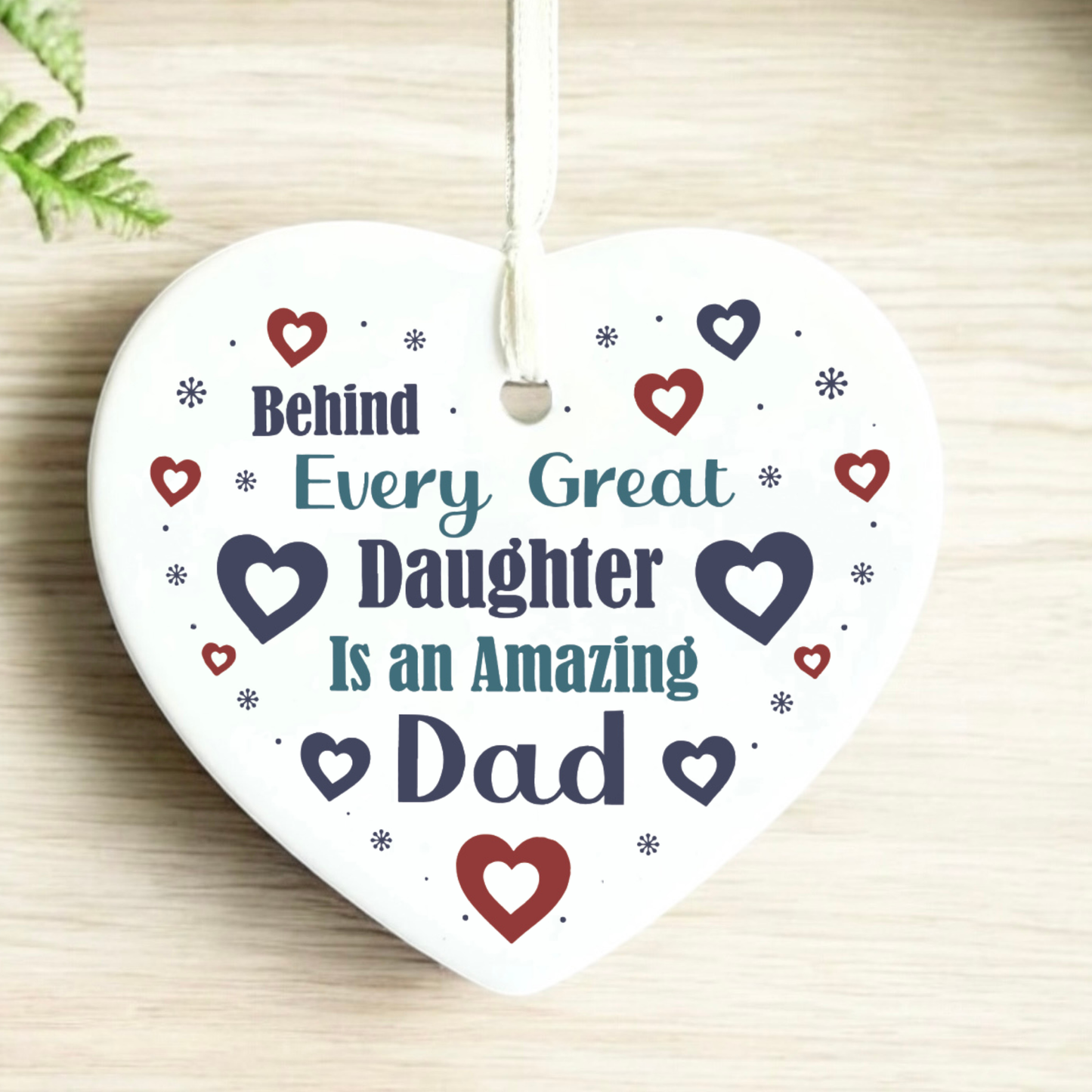 A close-up shot of a white ceramic heart-shaped hanging ornament displaying the text "Behind Every Great Daughter Is an Amazing Dad" surrounded by small red and navy hearts and tiny star patterns, complete with a white hanging ribbon.
