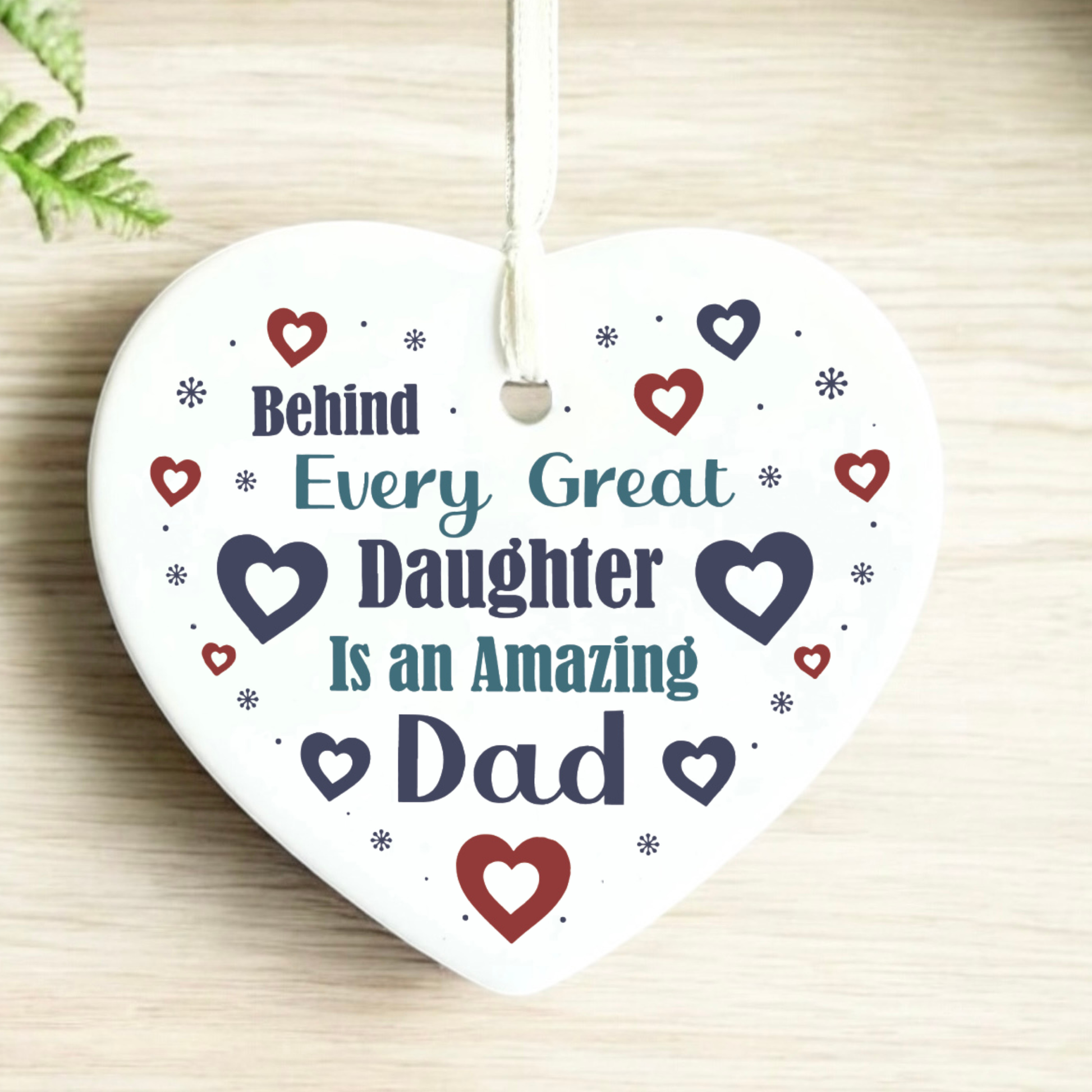 A close-up shot of a white ceramic heart-shaped hanging ornament displaying the text "Behind Every Great Daughter Is an Amazing Dad" surrounded by small red and navy hearts and tiny star patterns, complete with a white hanging ribbon.