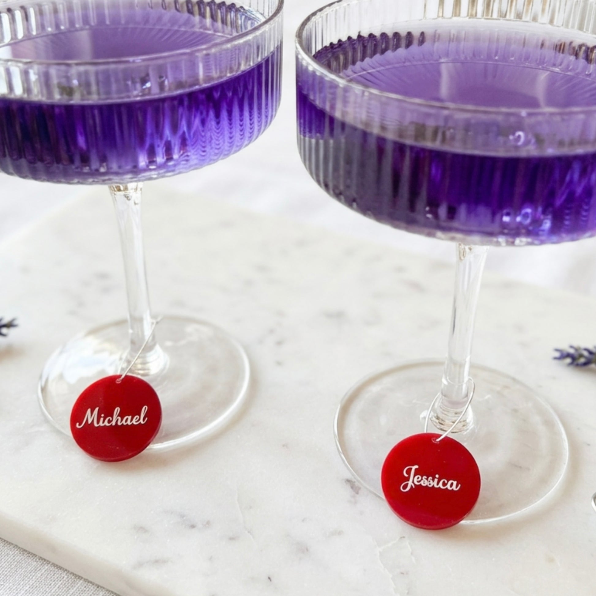 Two clear stemmed coupe glasses filled with purple liquid sit on a white marble surface, each with a small round red hanging tag engraved with a name: Michael on the left and Jessica on the right.