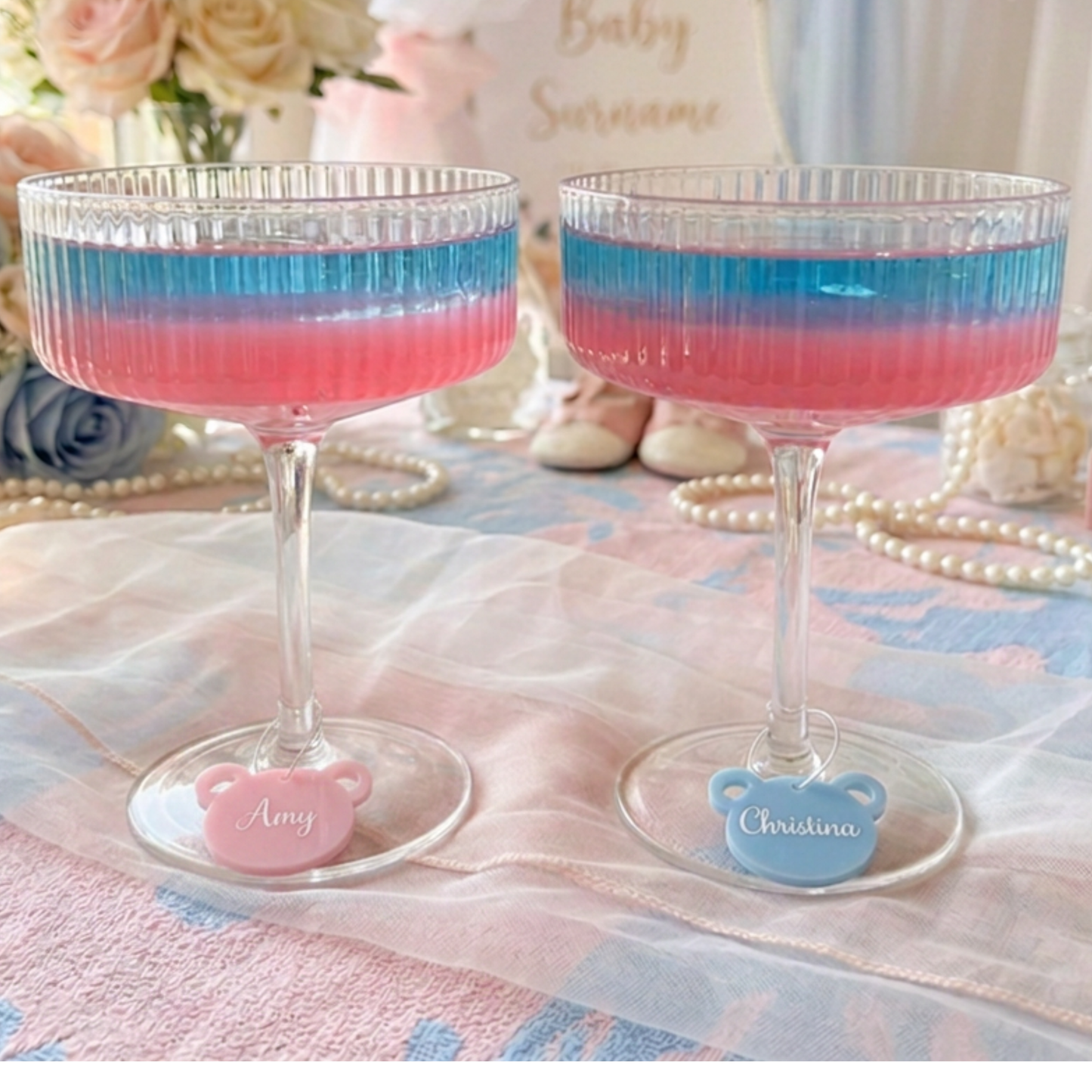 Two clear stemmed glass cups filled with pink and blue layered liquid, each with a small bear-shaped base charm personalized with a name, set on a pastel tablecloth with flowers and pearls in the background.