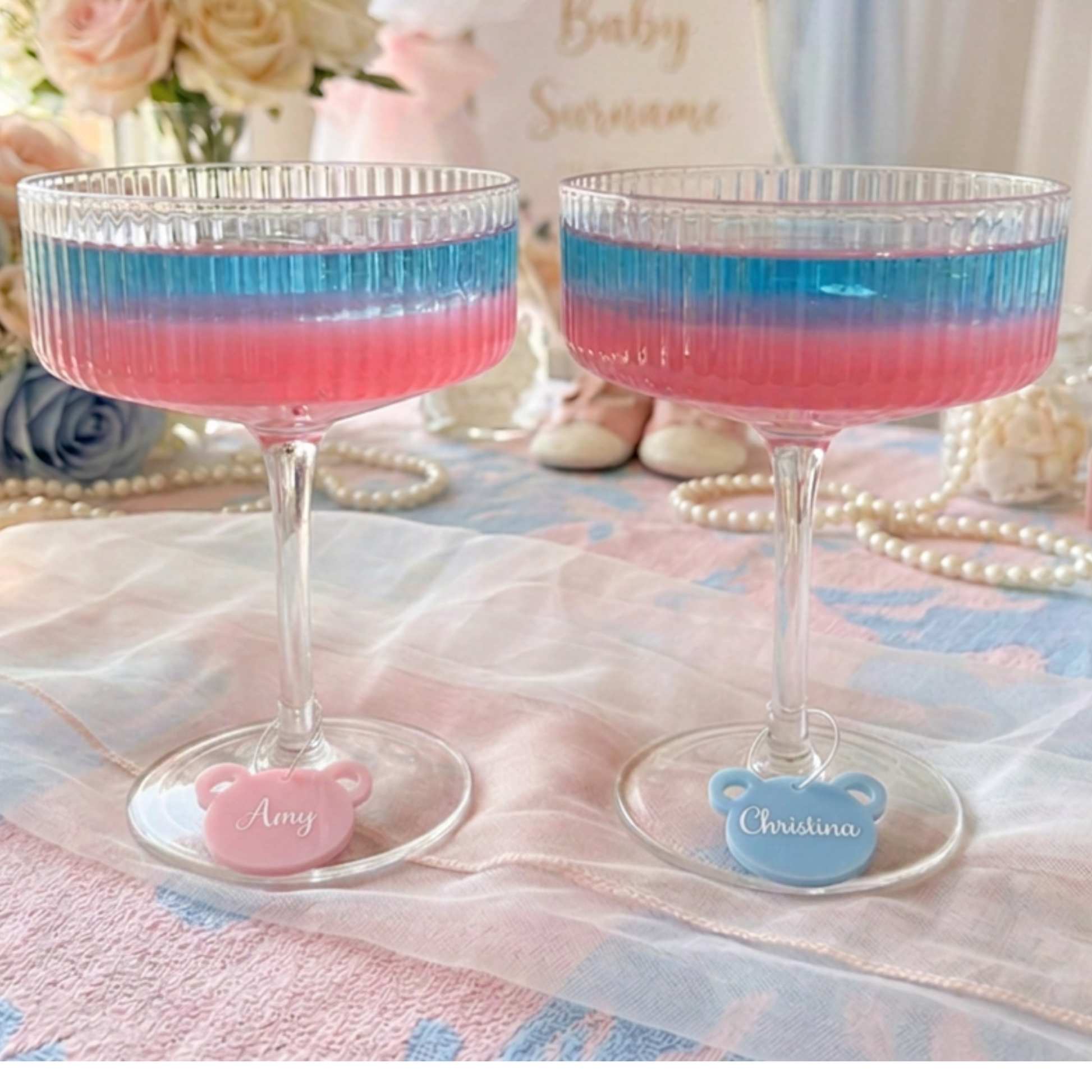 Two clear stemmed glass cups filled with pink and blue layered liquid, each with a small bear-shaped base charm personalized with a name, set on a pastel tablecloth with flowers and pearls in the background.