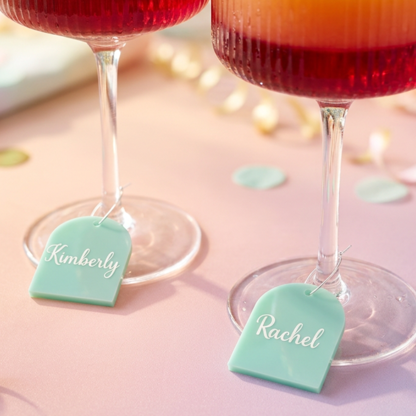 Two stemmed glasses filled with red drink on a pink surface, each with a mint green personalized tag hanging from the base. The tags are engraved with the names Kimberly and Rachel.