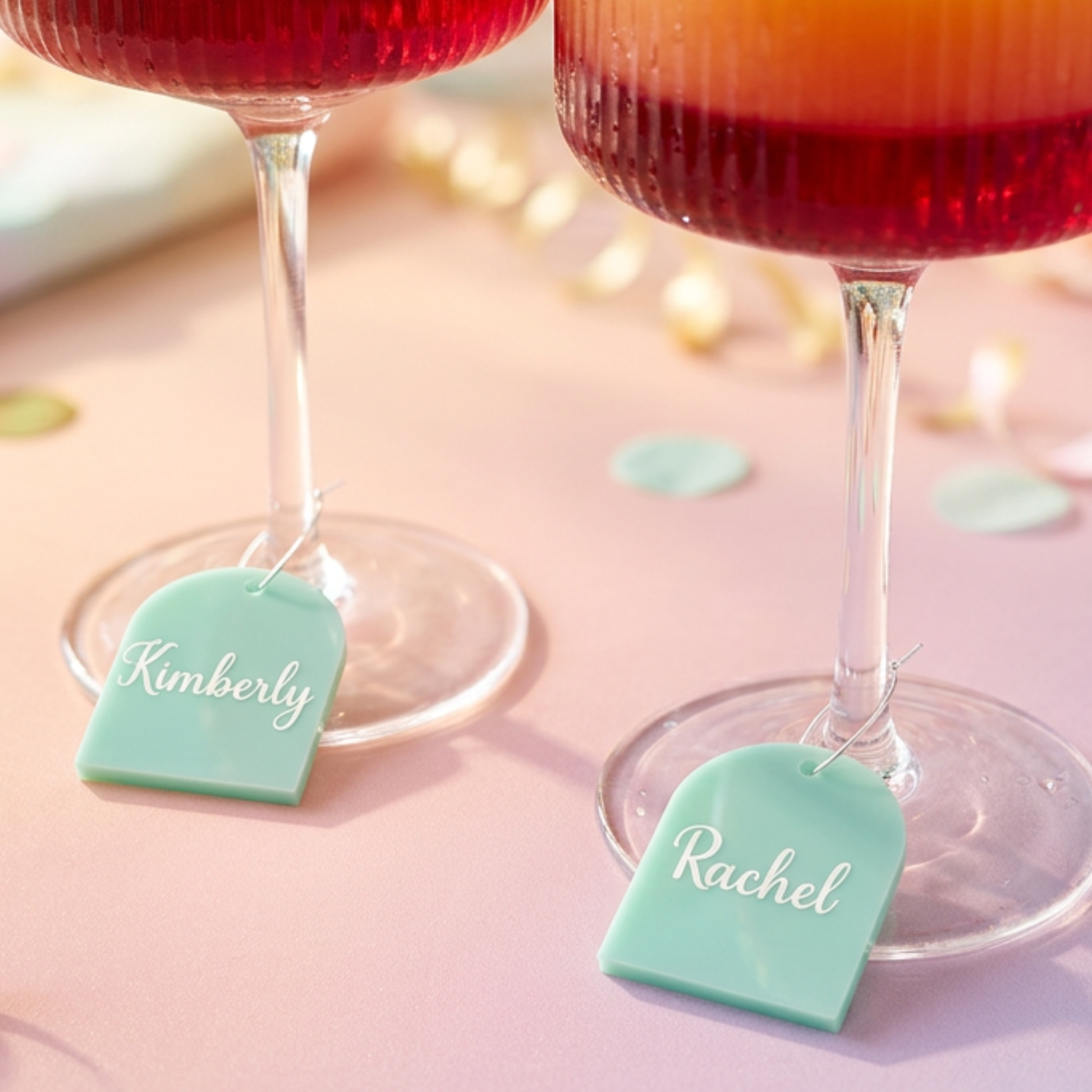 Two stemmed glasses filled with red drink on a pink surface, each with a mint green personalized tag hanging from the base. The tags are engraved with the names Kimberly and Rachel.