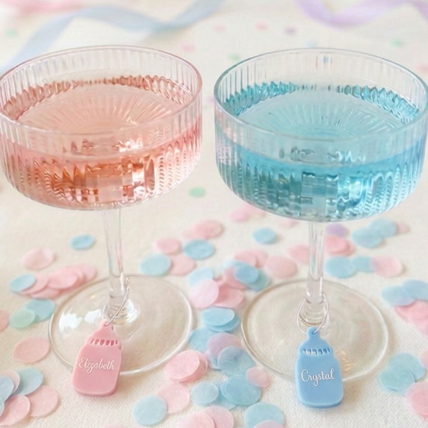 Two clear champagne coupe glasses filled with pink and blue liquid, each with a small hanging tag engraved with a name, set on a light surface with pastel confetti.