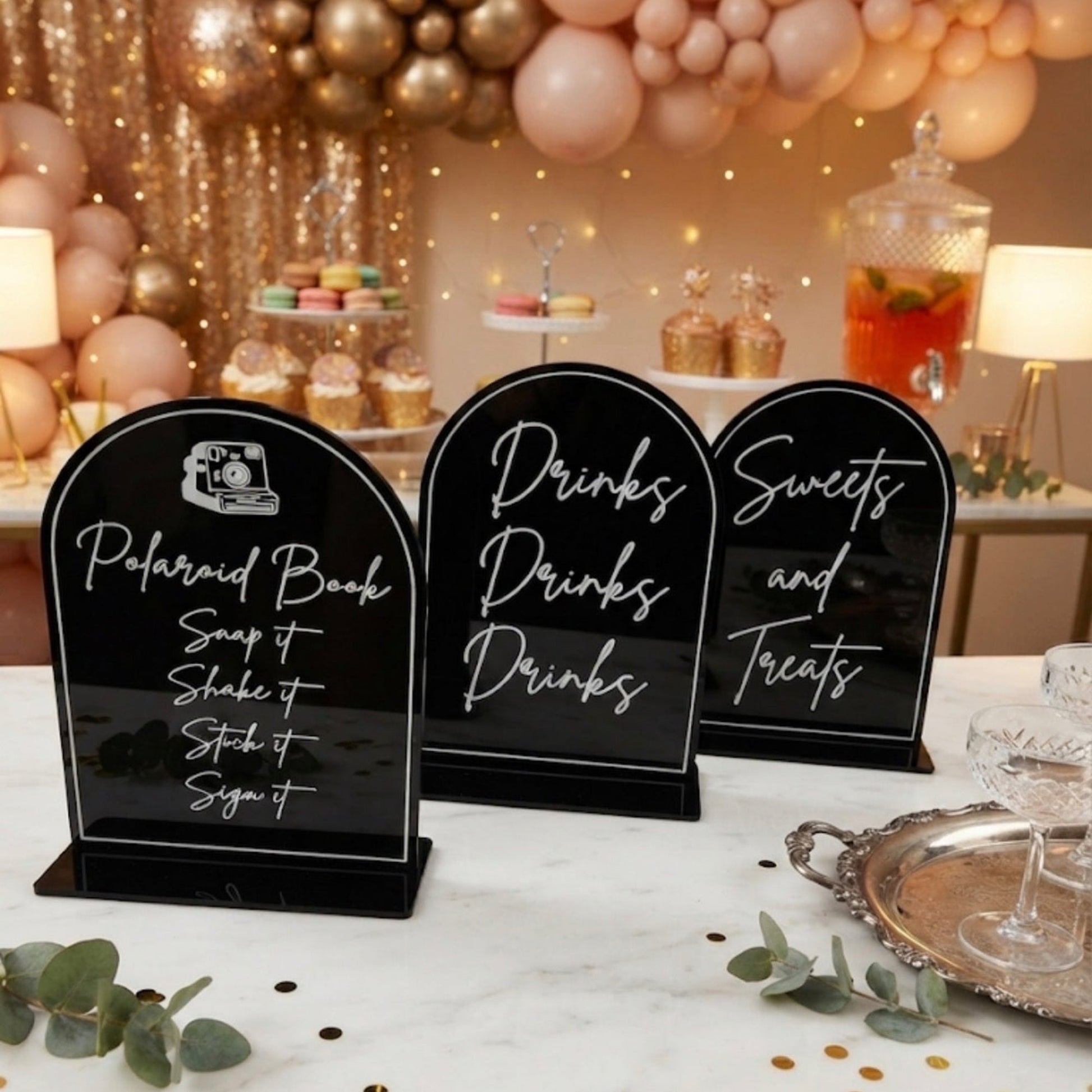 Three black acrylic sign holders with white script lettering are arranged on a white marble table at a decorated event. The signs read 'Polaroid Book', 'Drinks', and 'Sweets and treats'.