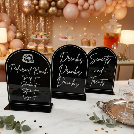 Three black acrylic sign holders with white script lettering are arranged on a white marble table at a decorated event. The signs read 'Polaroid Book', 'Drinks', and 'Sweets and treats'.