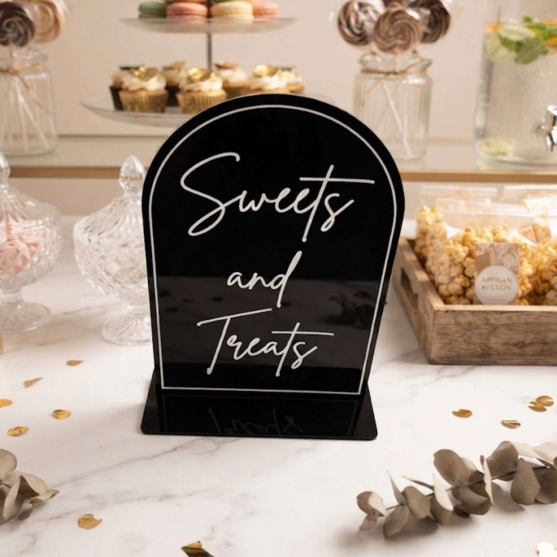 A black arched acrylic table sign with white script text reading 'Sweets and Treats,' displayed on a dessert table with cupcakes, macarons, lollipops, and popcorn in the background.