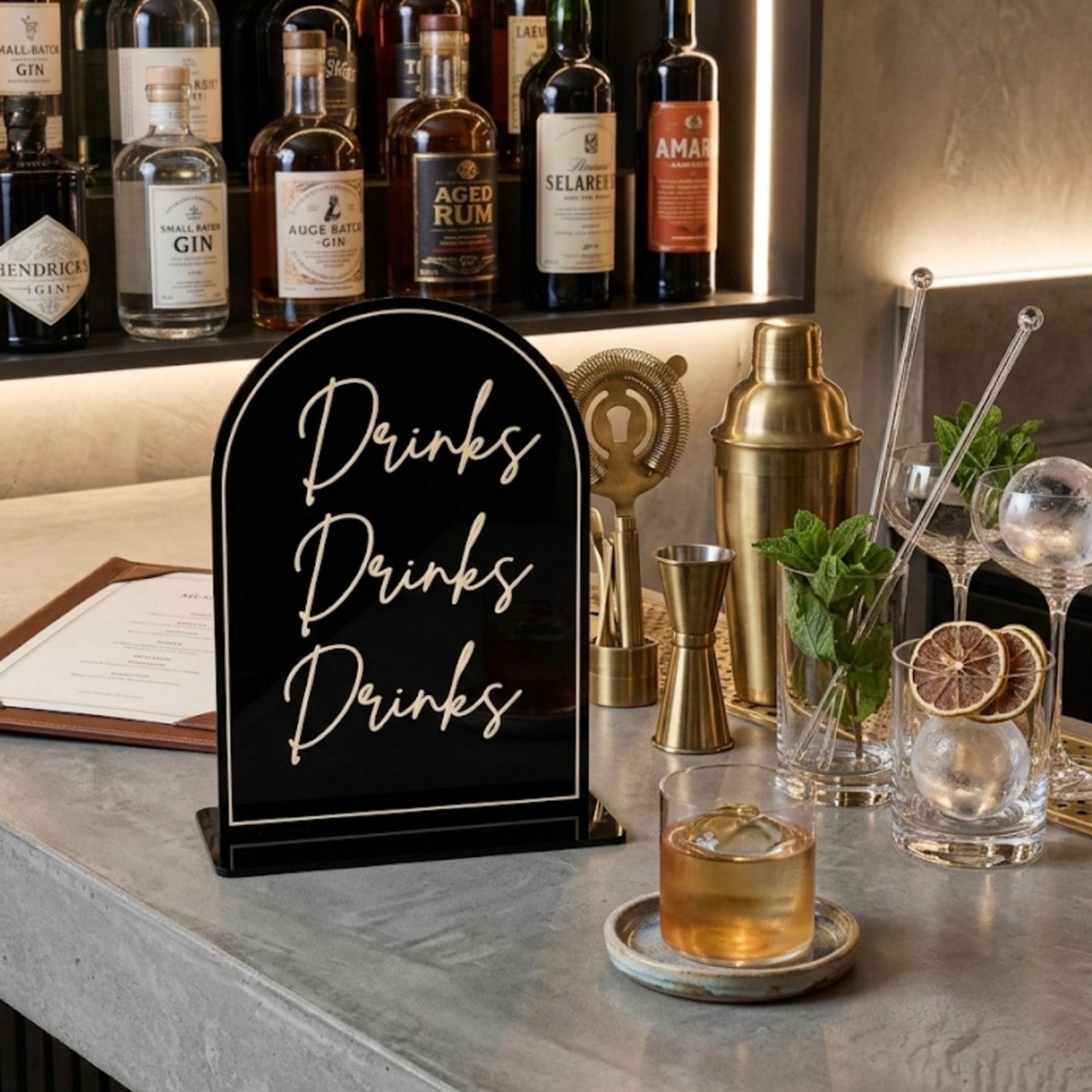 A black arched bar sign with white script reading 'Drinks Drinks Drinks' stands on a gray stone bar counter. In the background, liquor bottles are displayed on shelves with warm lighting, and bar tools including a gold shaker, straws, and a glass with an amber drink are arranged nearby.