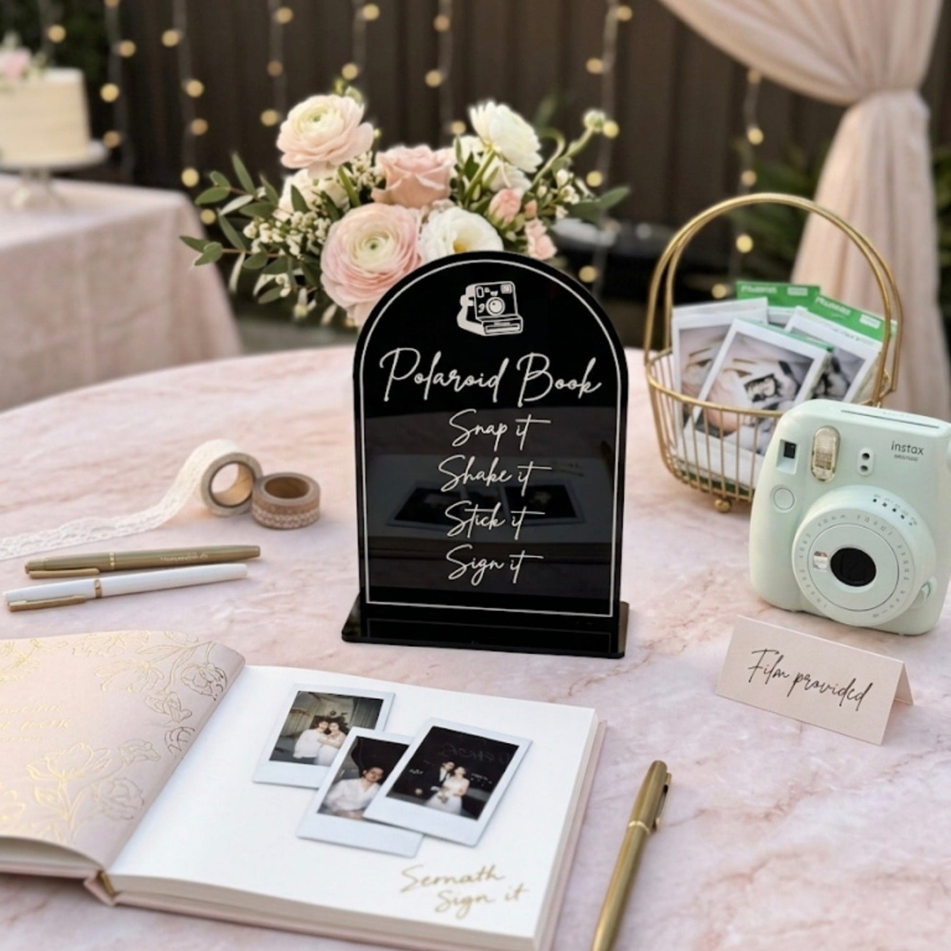 A styled wedding reception table setup featuring a black arched 'Purchased Book' sign, an open white guest book with printed photos, gold pens, a basket of programs, and a mint instant camera on a pale pink marble tablecloth.