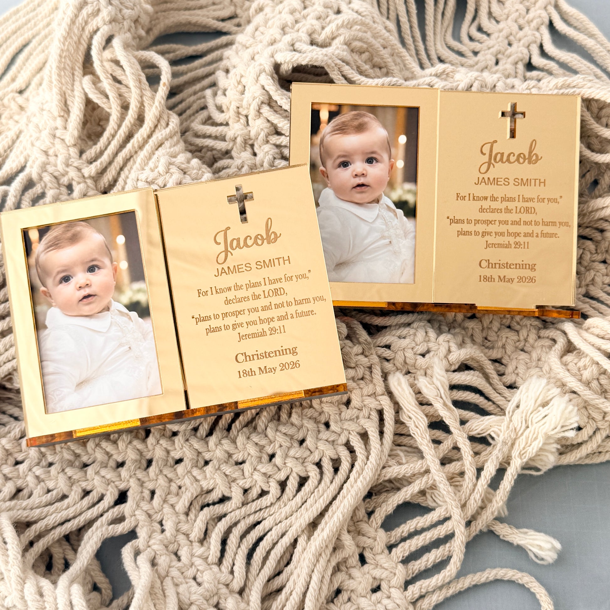 Personalized wooden baby christening photo frames displayed on a textured cream-colored knitted blanket. The set includes an open two-panel frame showing a baby photo and engraved text, and a closed three-panel frame with a small cross and scripture.