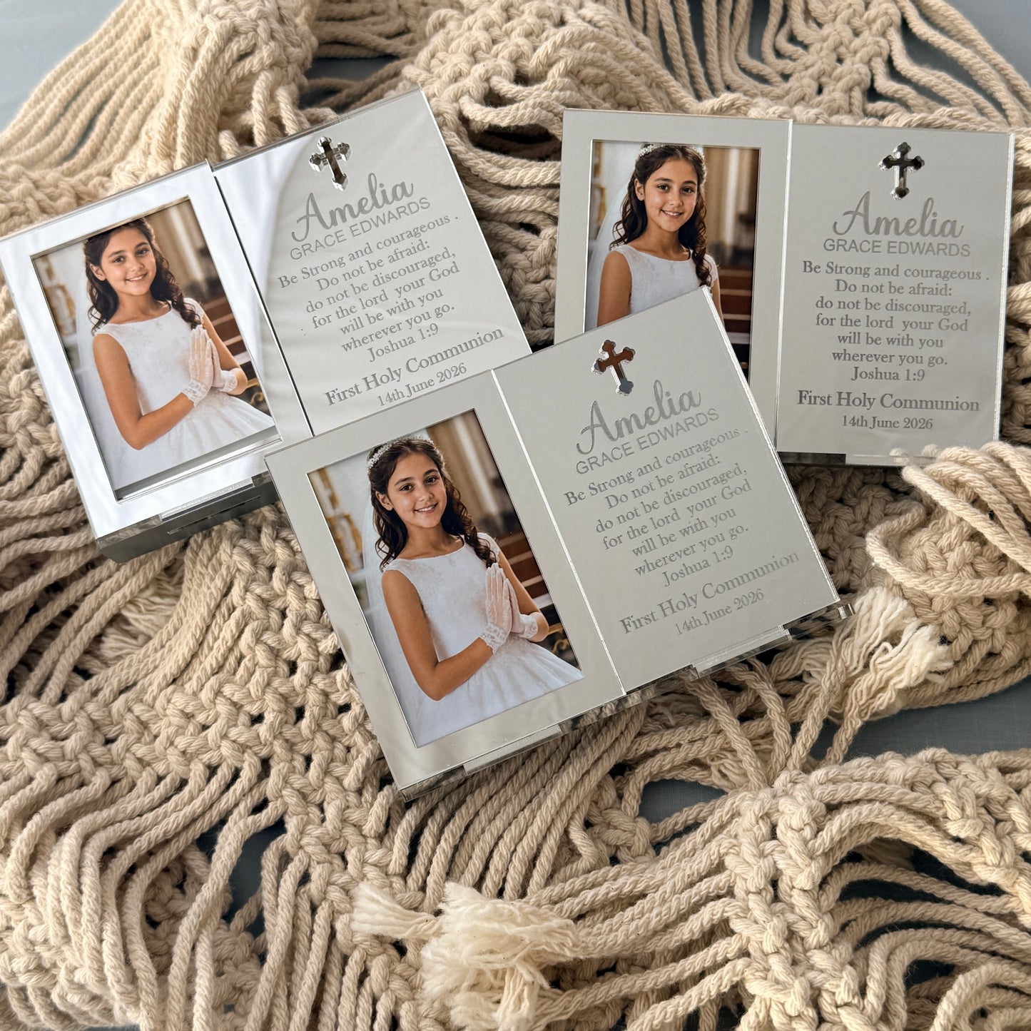 Four personalized first holy communion photo frames arranged on a beige woven rope blanket. The frames are silver with printed grace ordinances text and a photo of a young girl in a white dress, with one frame showing the back side with a cross and scripture.