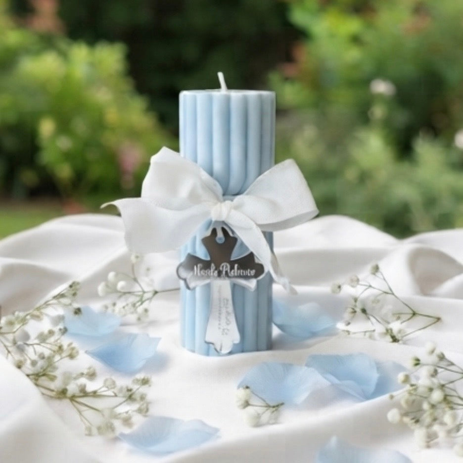 A light blue ribbed pillar candle tied with a white satin bow and a small hanging tag that reads 'Meads Palms', styled on white draped fabric with scattered pale blue petals and white baby's breath flowers, with a blurred green garden background.