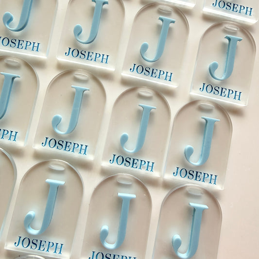 A close-up of a sheet of clear acrylic name tiles featuring a light blue raised letter 'J' and the name 'JOSEPH' printed below in blue, arranged in a grid on a white background.