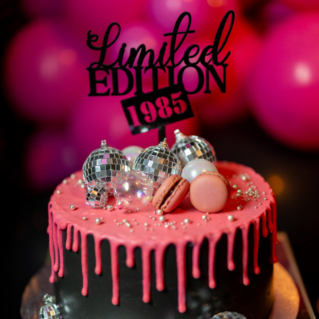 Decorative cake with pink icing, silver decorations, and 'Limited Edition 1985' topper against a blurred pink background.