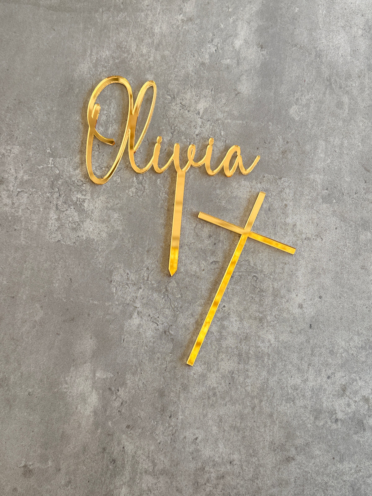 Gold metal personalized wall decor featuring the name 'Olivia' in cursive script and a cross, displayed on a textured gray concrete surface.