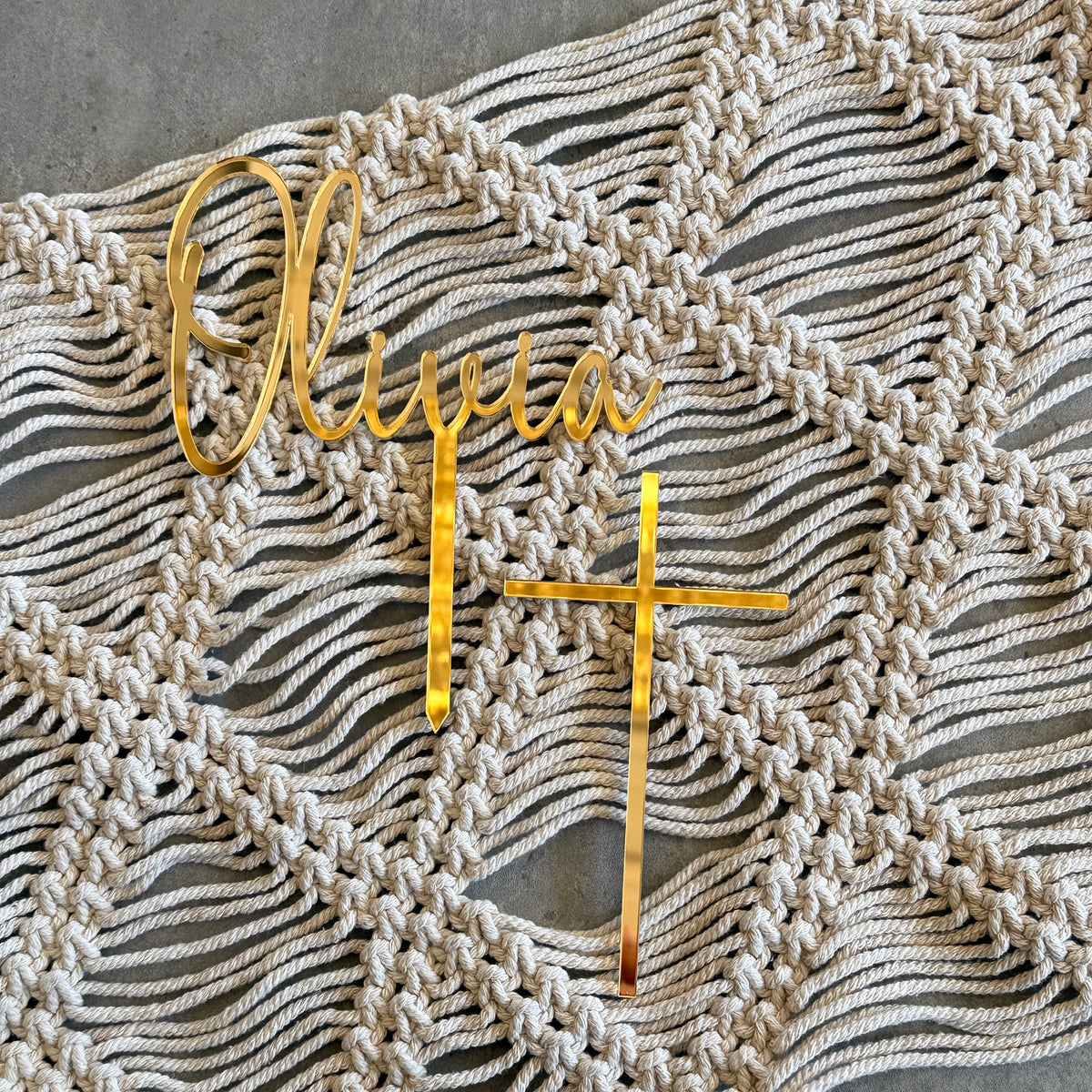 Gold metal personalized wall decor featuring the name 'Olivia' in cursive script, a vertical bar with a cross at the bottom, and a small gold cross, displayed on a cream macrame wall hanging over a gray concrete surface.
