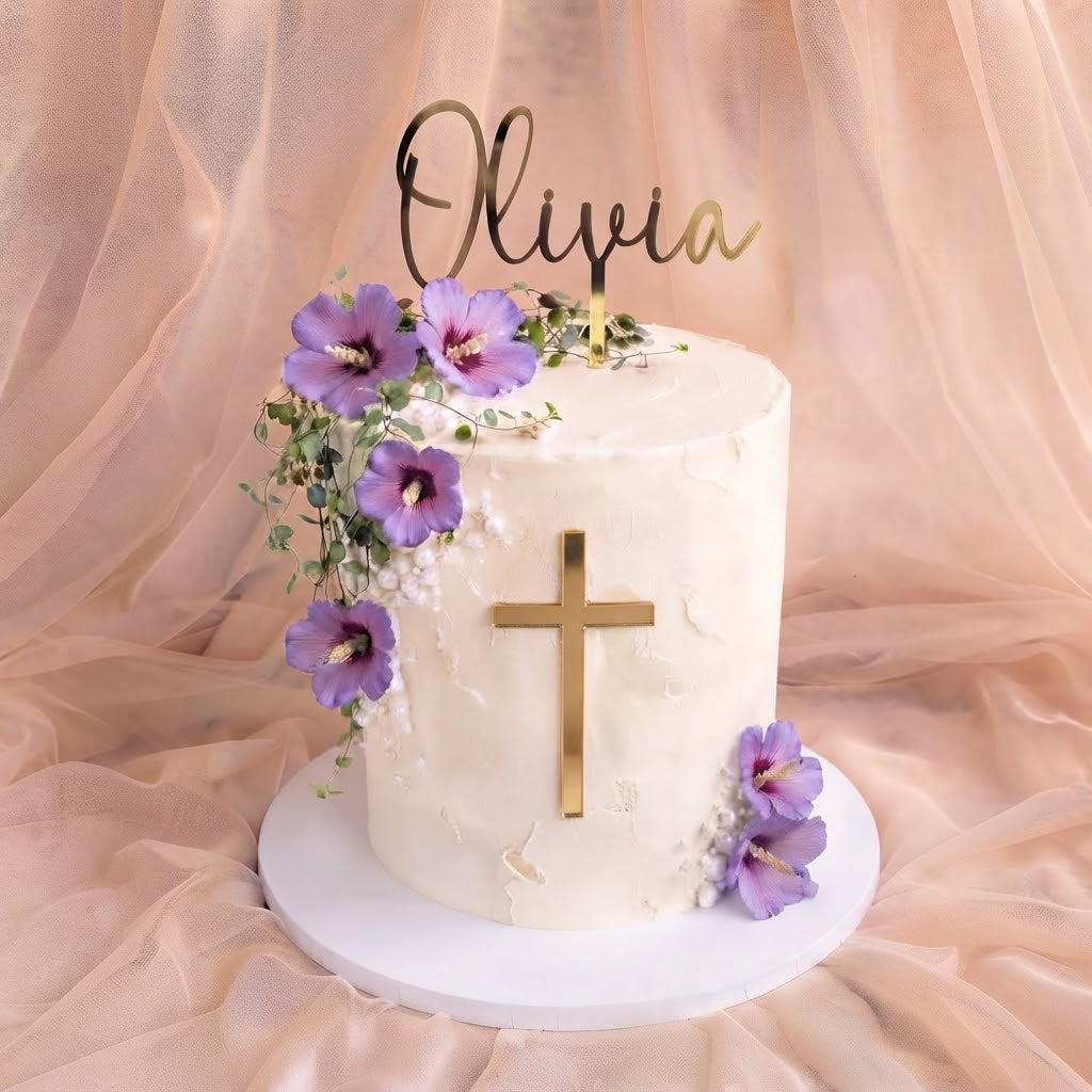 A frosted white cake with a gold cross on the front, purple flowers with green leaves, and a gold script topper reading 'Olivia', displayed on a white round cake stand against draped blush pink fabric.