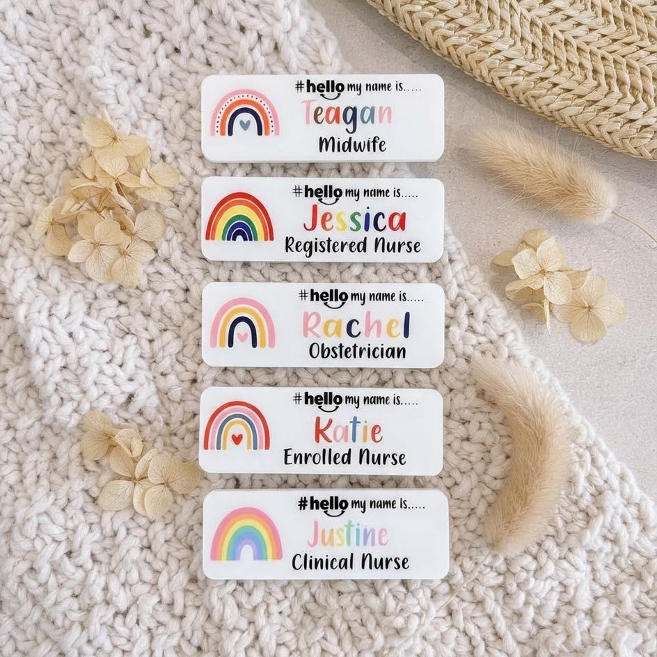 Five personalized white name tags with colorful rainbow graphics are arranged on a cream knit blanket with dried flowers and a woven straw hat nearby. The tags include sample names and titles such as Midwife, Registered Nurse, Obstetrician, Enrolled Nurse, and Clinical Nurse.