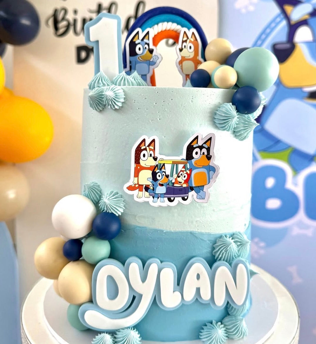 A two-tier light blue and dark blue ombre buttercream birthday cake decorated with Bluey character details, featuring a custom 14cm wide double-layer acrylic name fropper reading Dylan on the front, a matching number 1 topper on top, and coordinating blue, white, and tan balloon balls.
