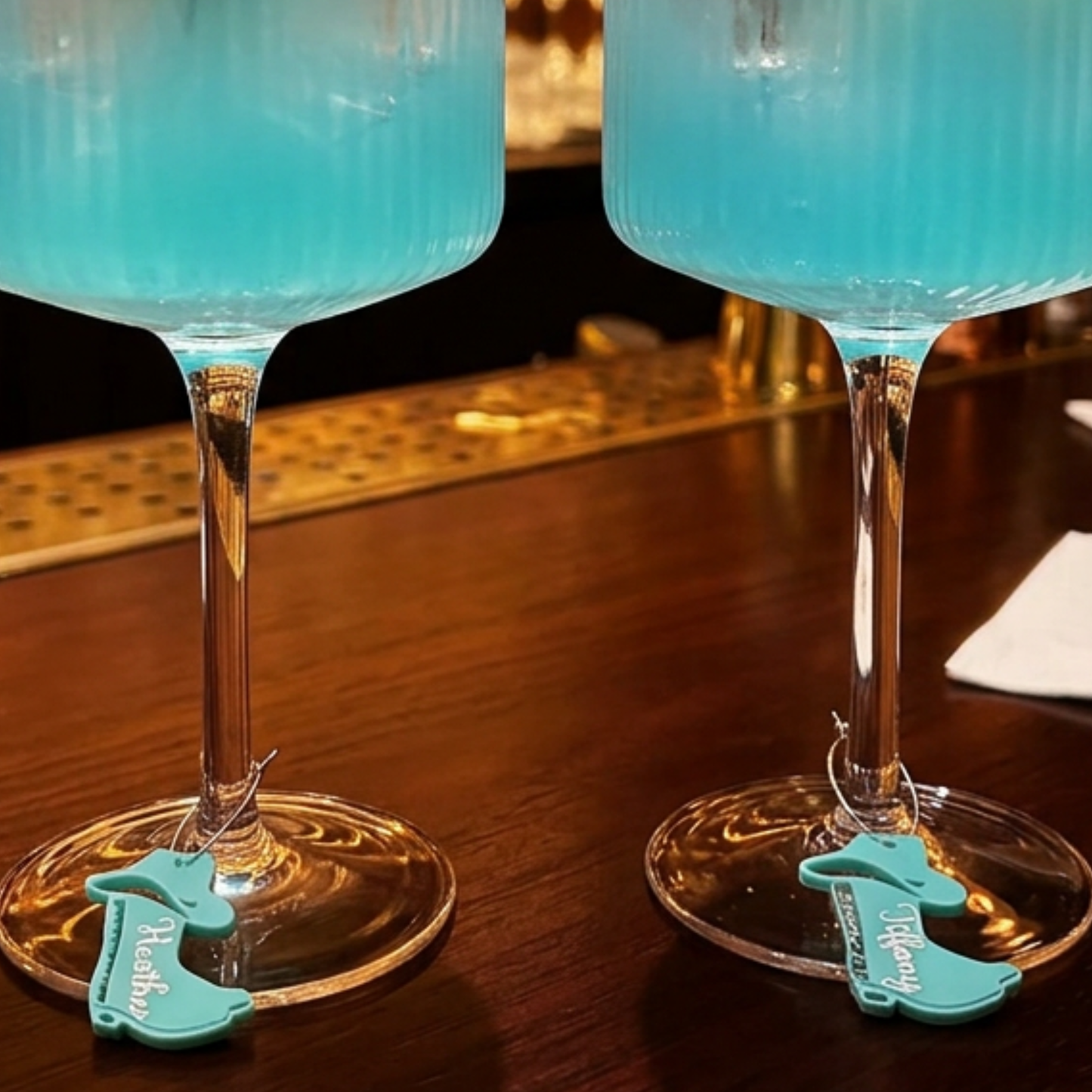 Two clear stemmed glasses filled with bright turquoise-blue liquid on a dark wooden bar counter, each with a small turquoise hang tag attached near the base.