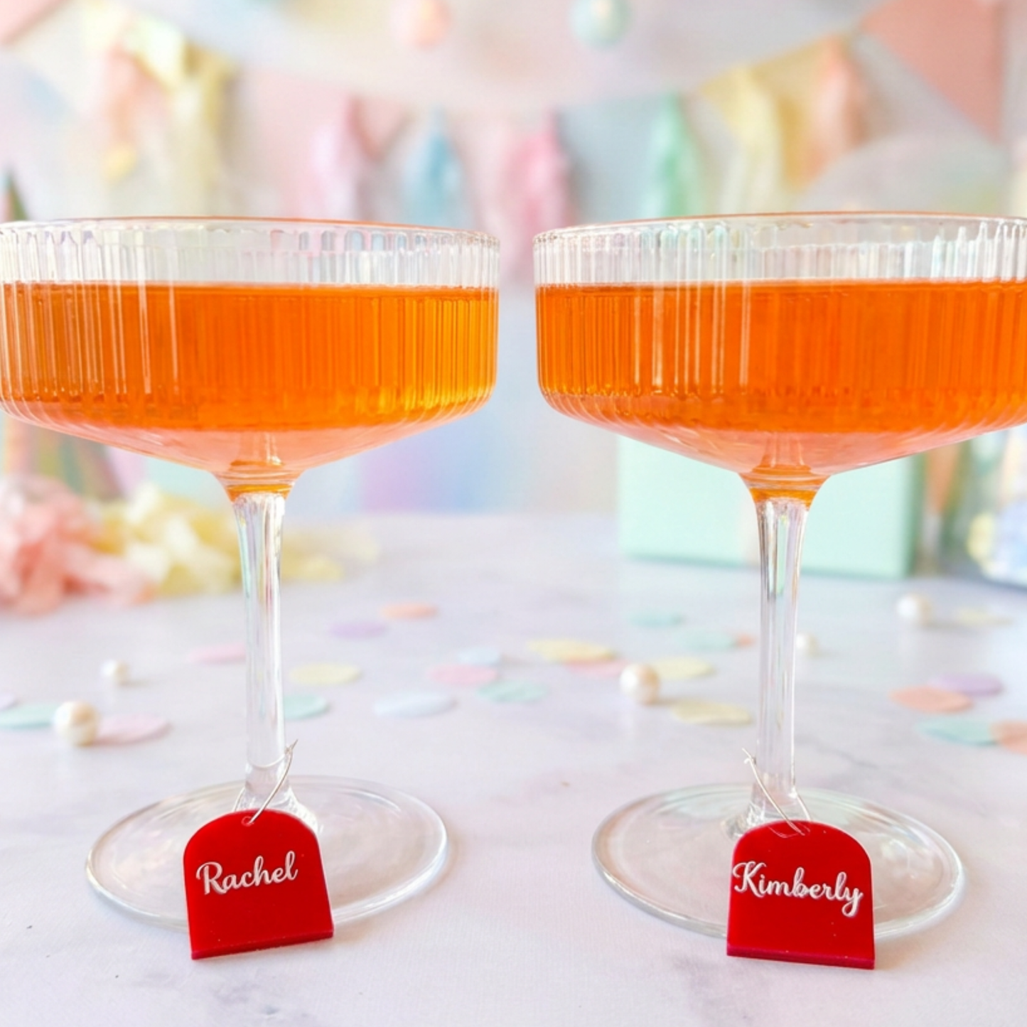 Two clear stemmed coupe glasses filled with orange liquid, each with a small red hanging tag labeled Rachel and Kimberly, set on a light surface with pastel confetti and a soft blurred party background.