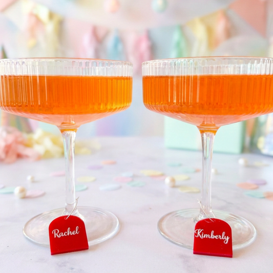 Two clear stemmed coupe glasses filled with orange liquid, each with a small red hanging tag labeled Rachel and Kimberly, set on a light surface with pastel confetti and a soft blurred party background.