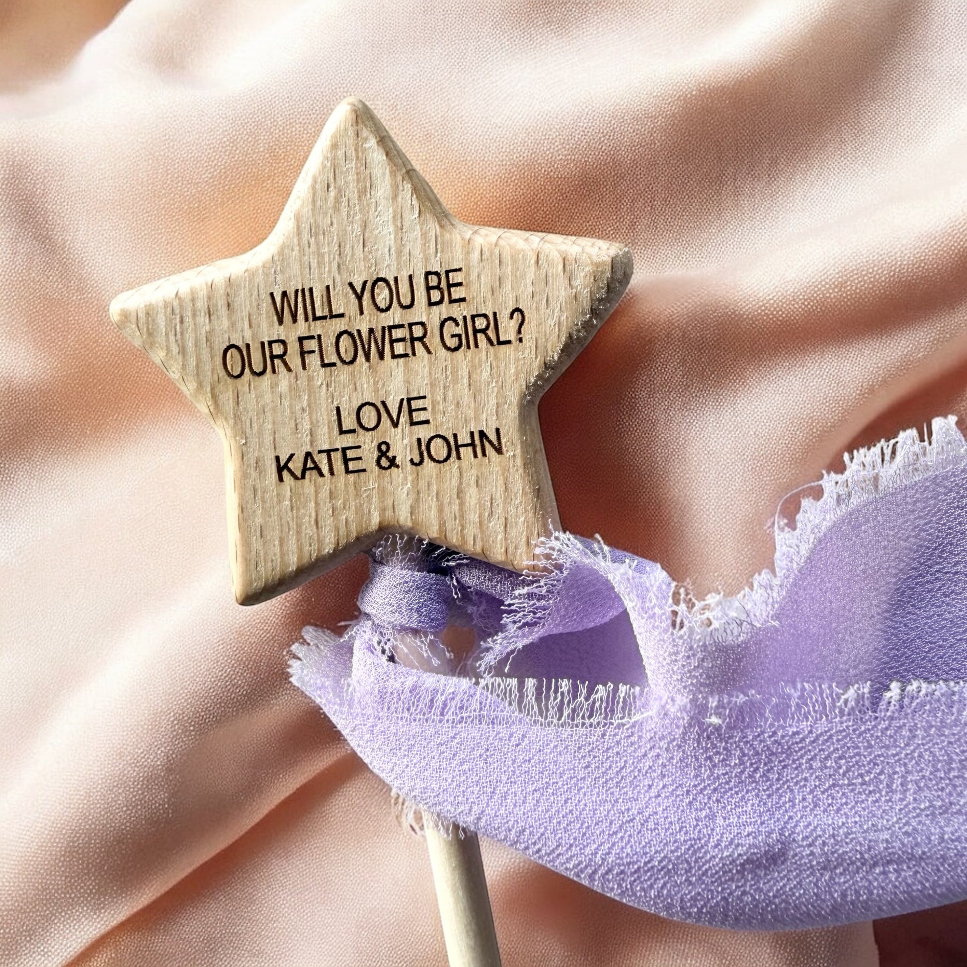 Star-shaped wooden prop with 'Will you be our flower girl?' message held by a person wearing a purple dress.