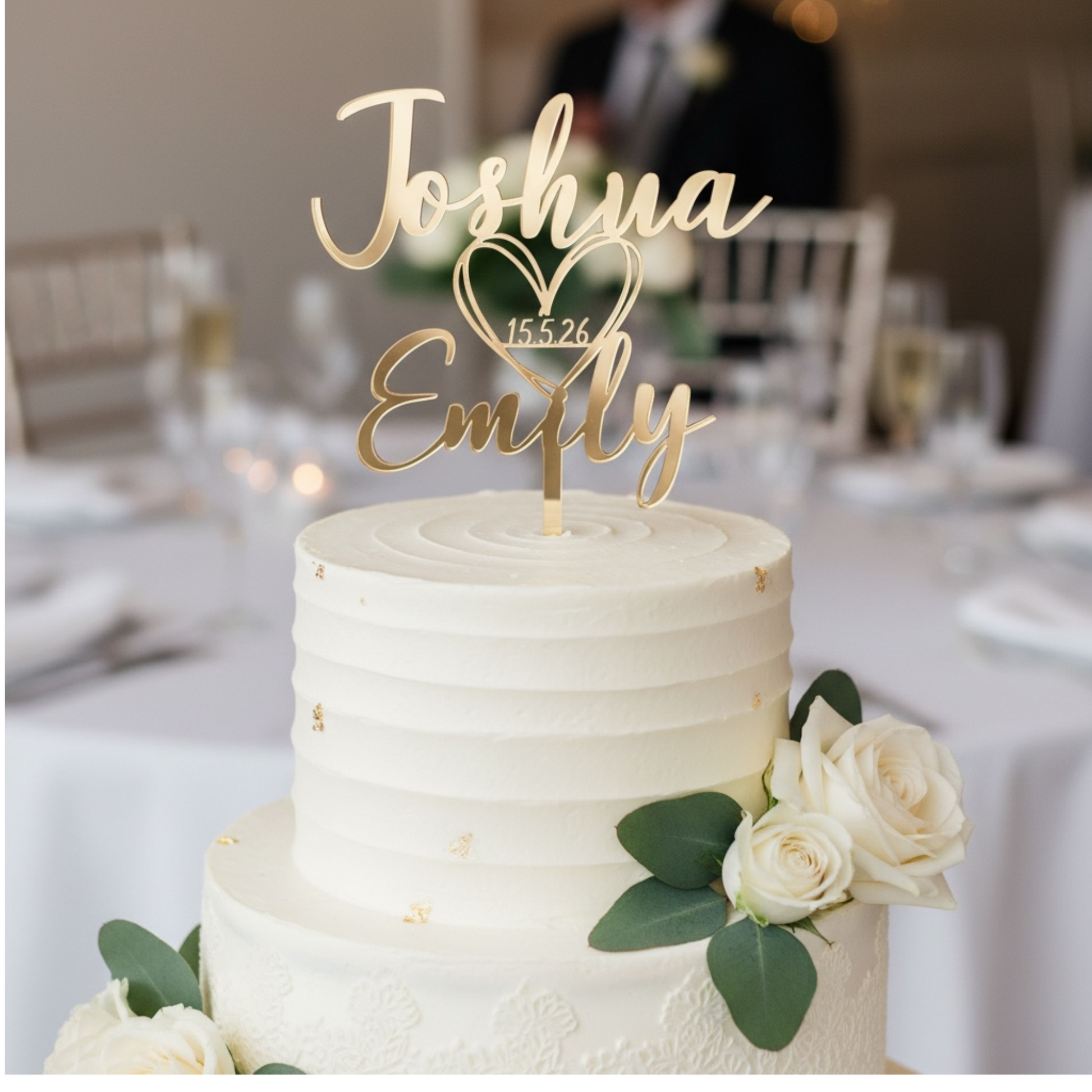 A two-tier white wedding cake with gold leaf accents and white roses, topped with a gold custom name cake topper reading 'Joshua Emily' with a heart and the date '15.5.