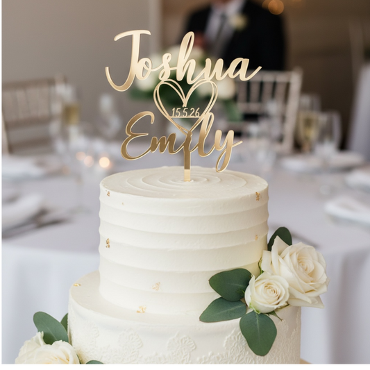 A two-tier white wedding cake with gold leaf accents and white roses, topped with a gold custom name cake topper reading 'Joshua Emily' with a heart and the date '15.5.