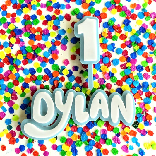 Double layer light blue and white acrylic cake topper set featuring a 14cm wide name fropper reading Dylan and a matching number 1 topper, displayed on a vibrant multicolored confetti background.