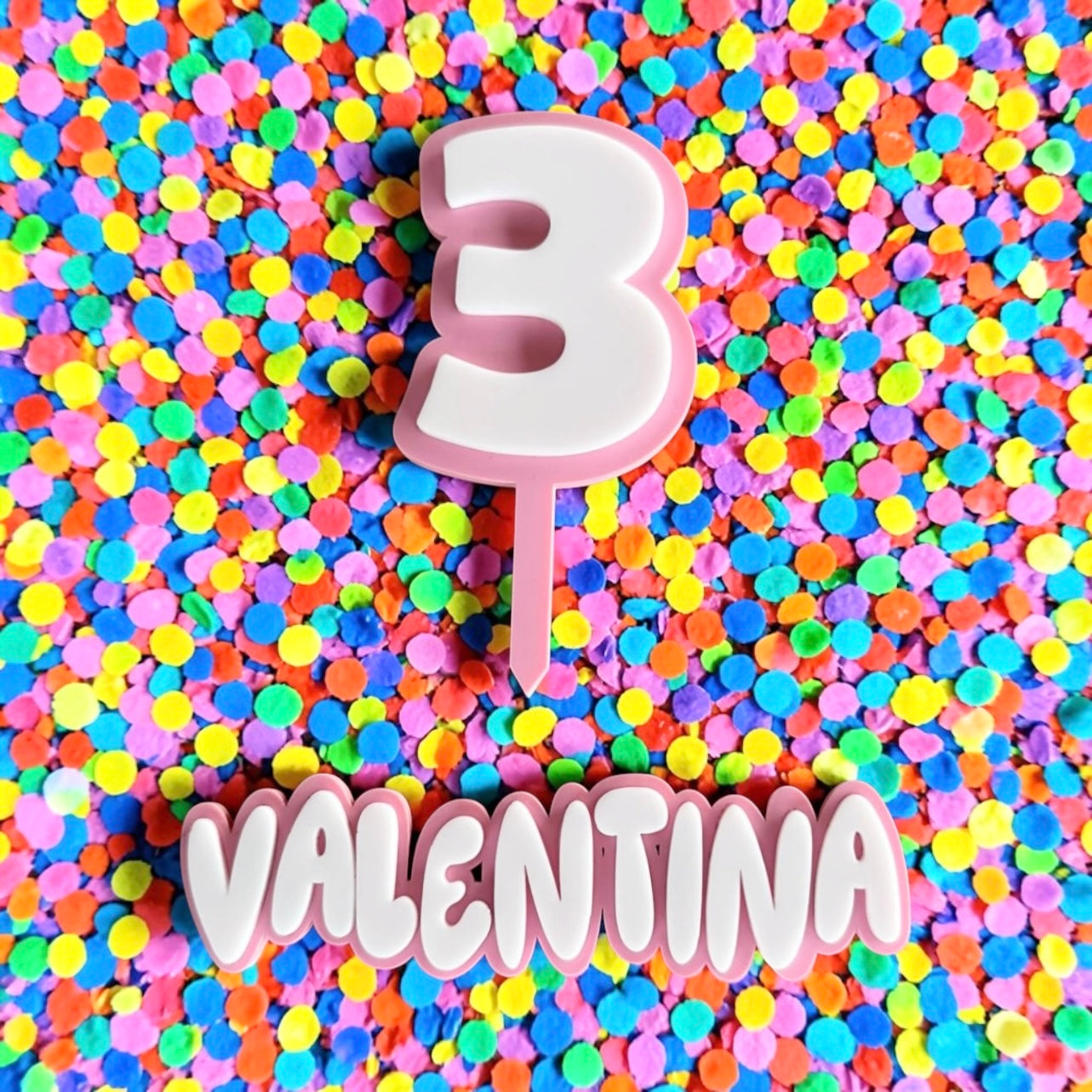 Custom pastel pink and white double layer acrylic cake decoration set with a script name fropper reading Valentina and a matching number 3 topper on a stake, over a bright colorful confetti backdrop.