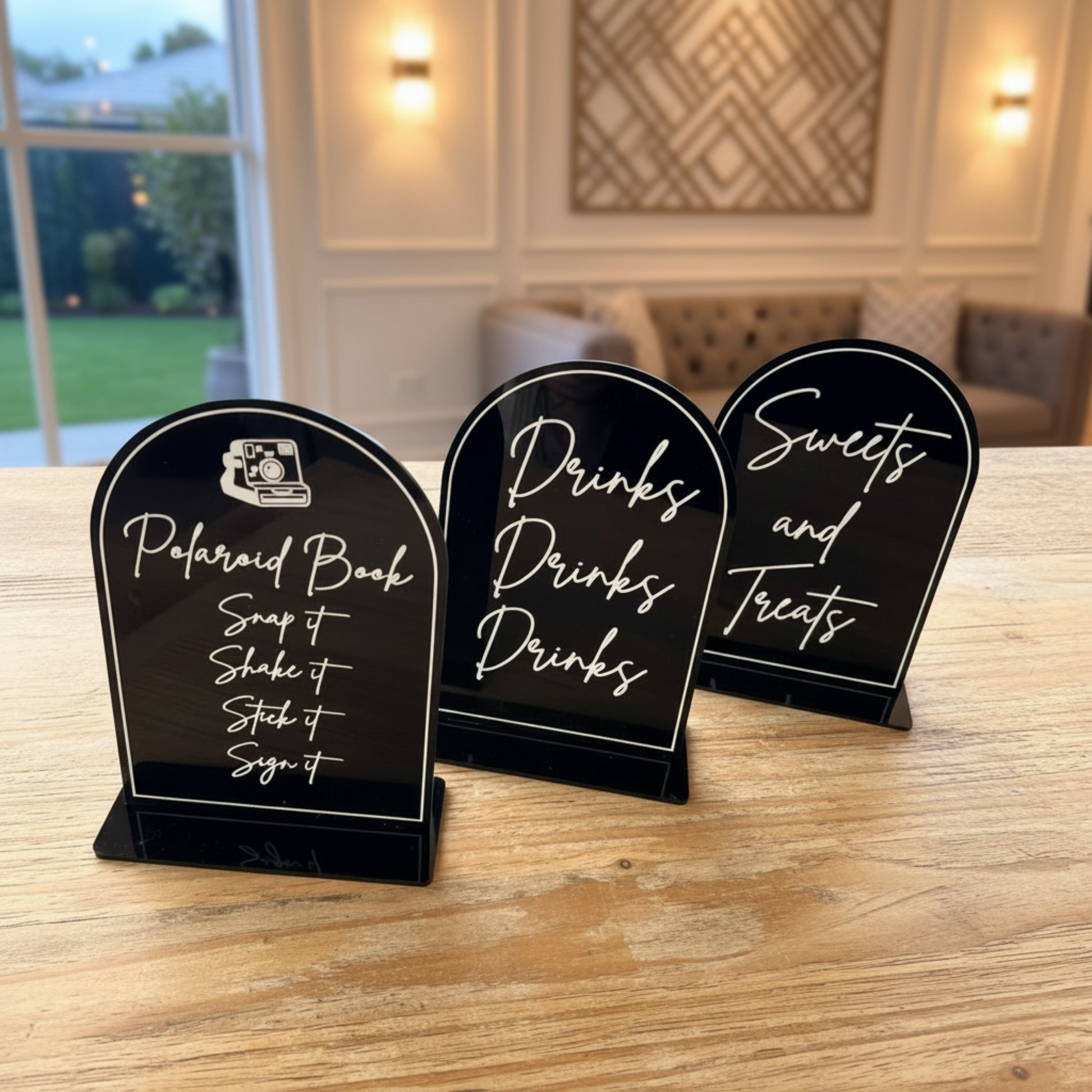 Three black tombstone-style sign holders on a wooden table, each with white script text reading 'Polaroid Book,' 'Drinks,' and 'Sweets and Treats,' in a softly lit indoor setting.