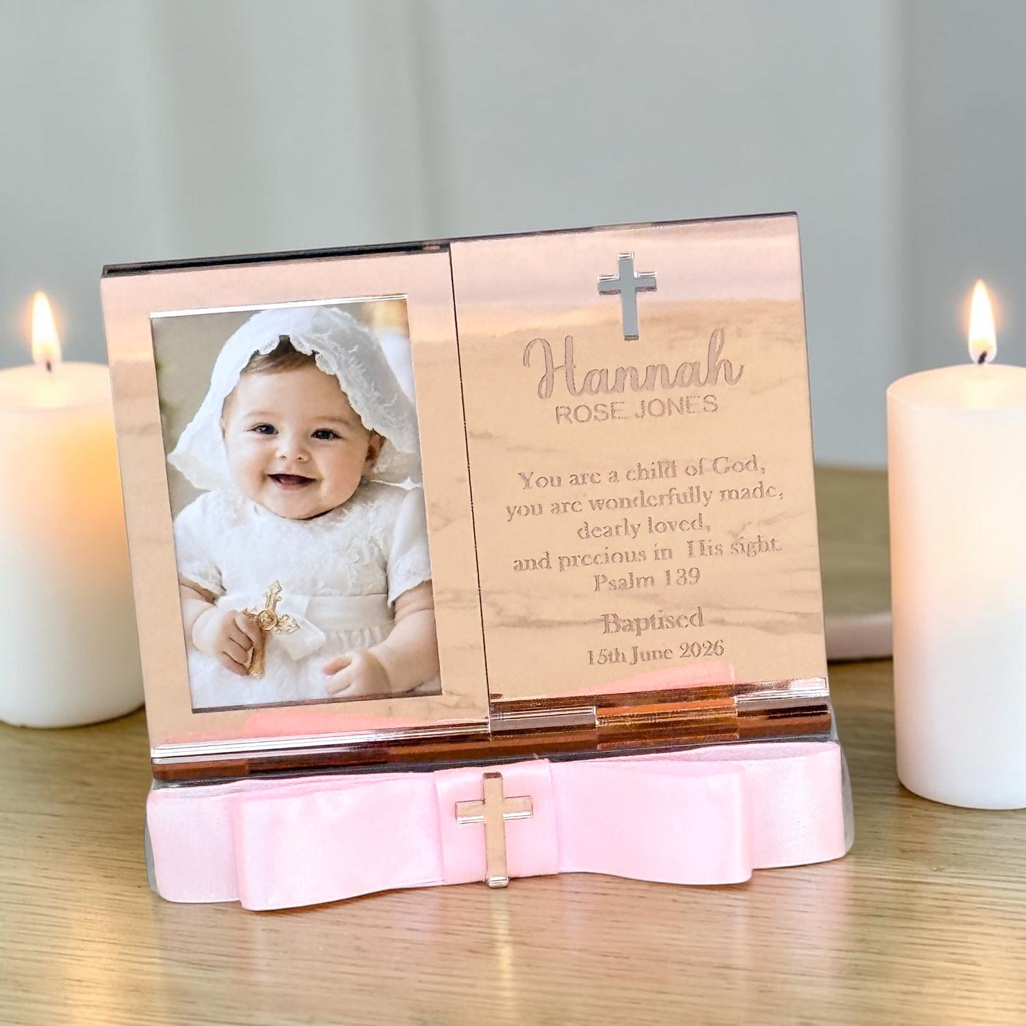 Personalized wooden baby baptism photo frame with a pink satin bow base, shown on a tabletop with two lit white candles. The frame holds a photo of a baby in a white outfit and has engraved text and a cross on the wooden panel.