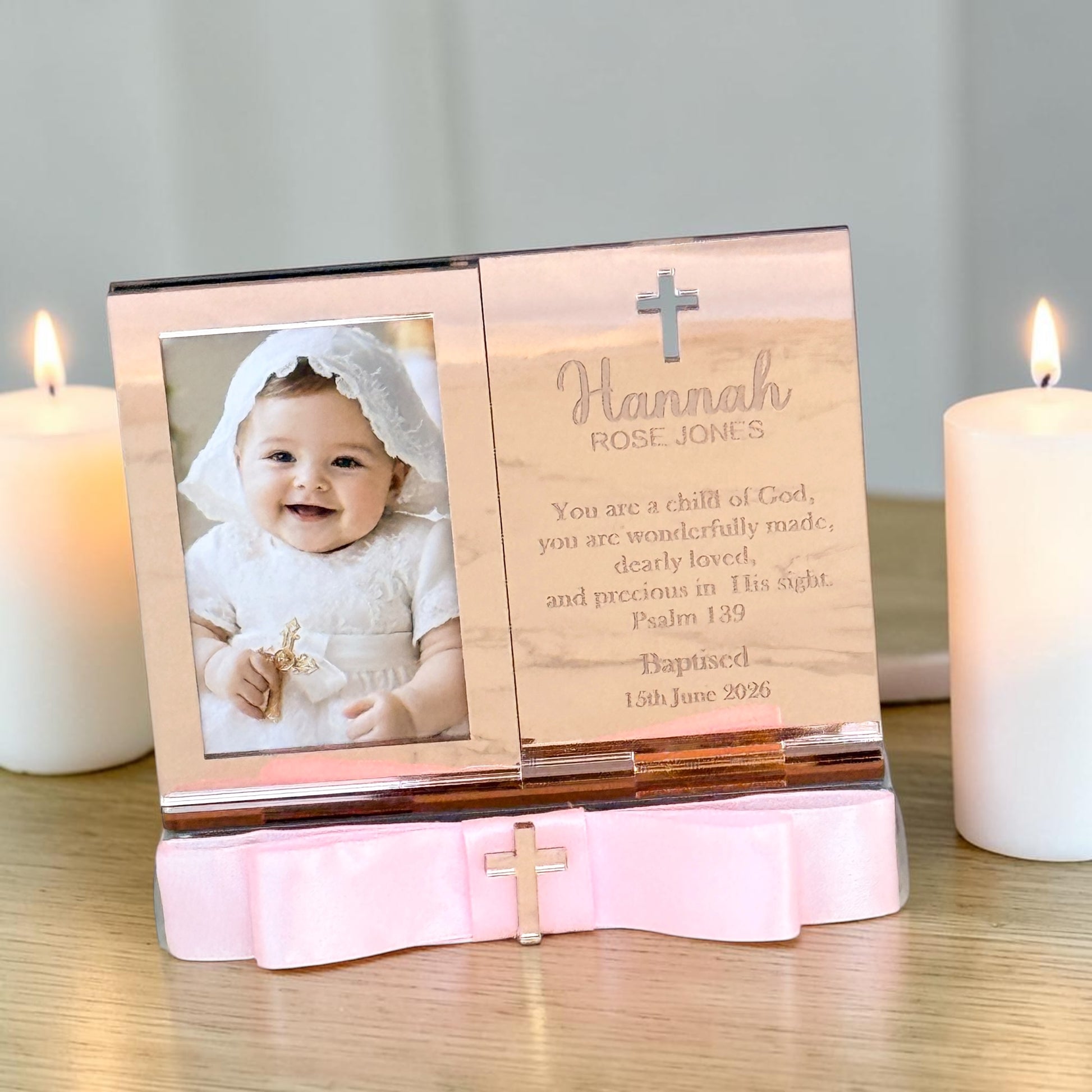 Personalized wooden baby baptism photo frame with a pink satin bow base, shown on a tabletop with two lit white candles. The frame holds a photo of a baby in a white outfit and has engraved text and a cross on the wooden panel.