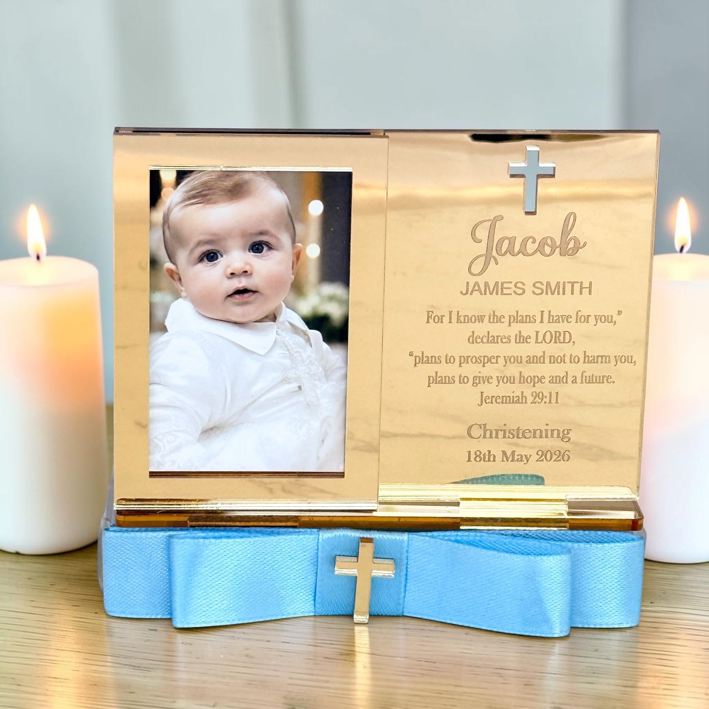 A personalized wooden baby christening keepsake box with a photo of a baby on the left and engraved text on the right, resting on a blue satin ribbon with a small gold cross, flanked by two lit white candles.