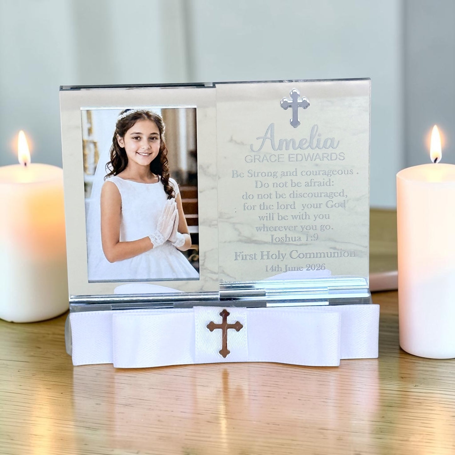 Personalized First Holy Communion photo frame with a white ribbon base, displayed on a wooden table between two lit candles. The left side shows a photo of a young girl in a white communion dress, and the right side has a marble-textured background with the name 'Amelia Grace Edwards' and a prayer printed in elegant script and serif fonts.