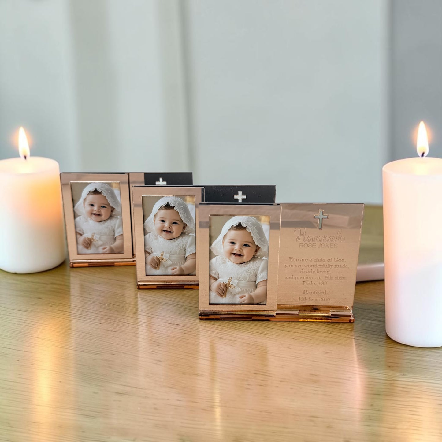 Three small rectangular photo frames with metallic rose-gold-toned borders are arranged on a wooden surface between two lit white candles. The center frame is open, showing a baby photo and a printed message card inside, while the side frames are closed.