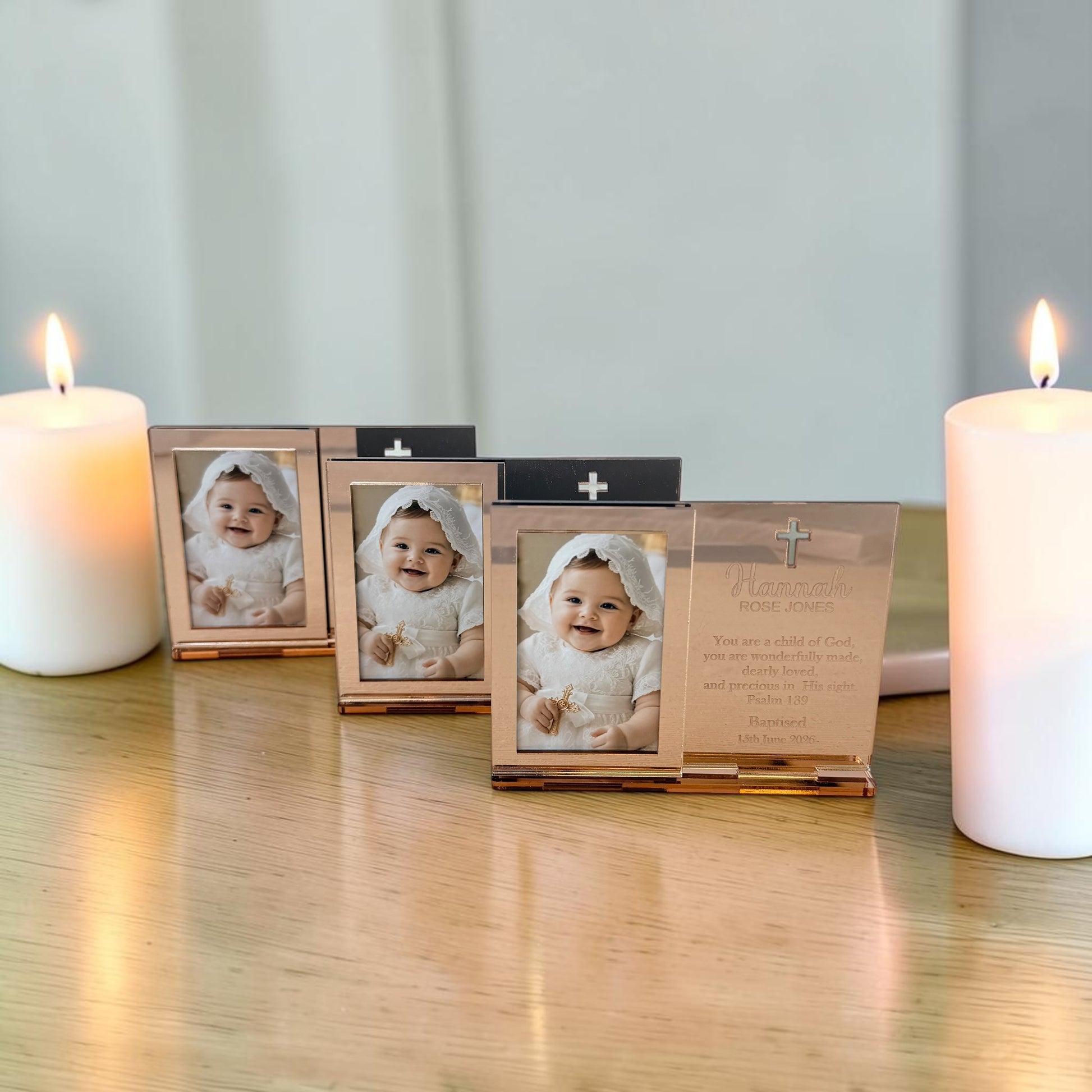 Three small rectangular photo frames with metallic rose-gold-toned borders are arranged on a wooden surface between two lit white candles. The center frame is open, showing a baby photo and a printed message card inside, while the side frames are closed.