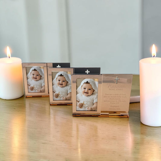 Three small rectangular photo frames with metallic rose-gold-toned borders are arranged on a wooden surface between two lit white candles. The center frame is open, showing a baby photo and a printed message card inside, while the side frames are closed.