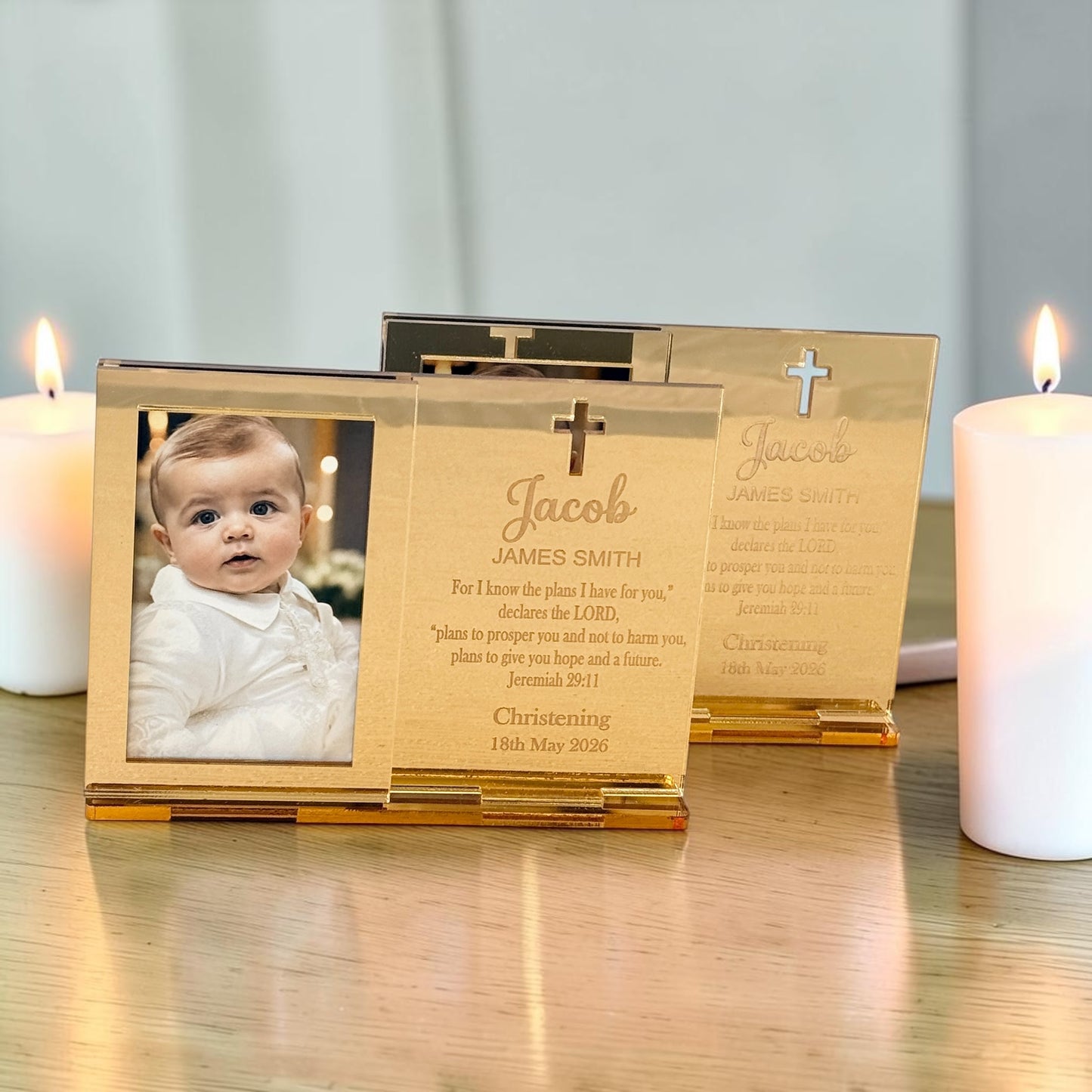 Personalized gold-toned wooden photo frames displayed on a wooden surface with lit white candles. The front frame shows a baby photo, while the two back frames are engraved with the name 'Jacob', a cross, and a Christian remembrance message.