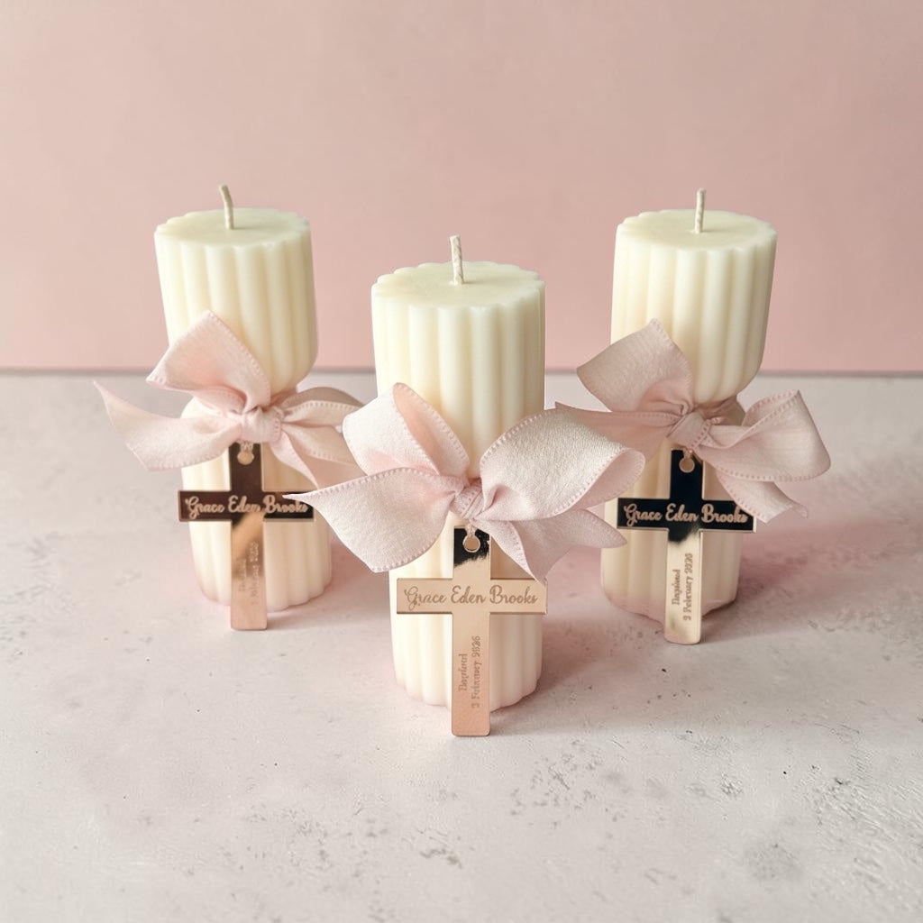 Three cream-colored ribbed pillar candles tied with pale pink satin ribbons and small wooden crosses, displayed on a light surface against a soft pink background.