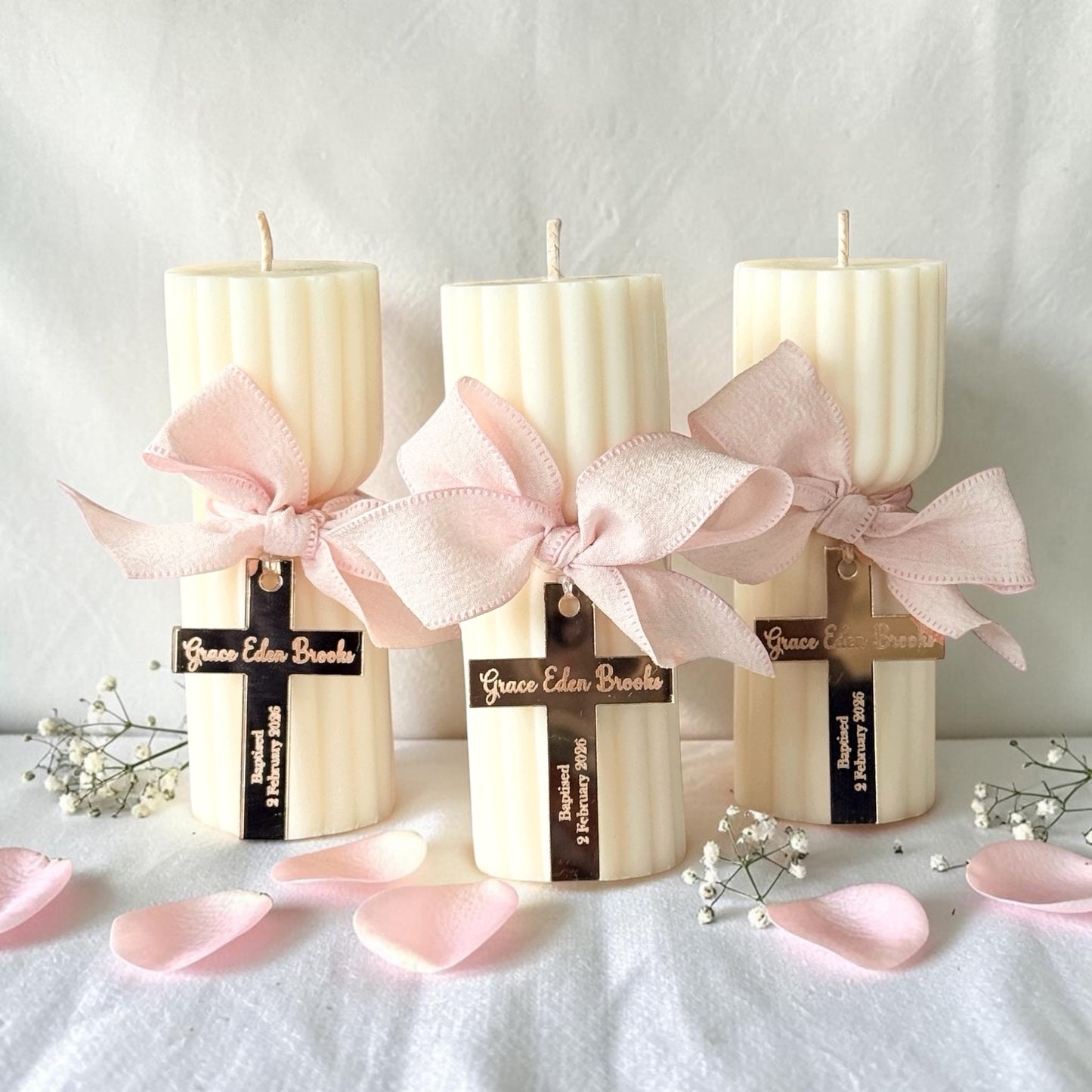 Three cream-colored ribbed pillar candles tied with large pale pink satin bows and black ribbon labels, arranged on a white fabric surface with scattered pink petals and small white flowers.