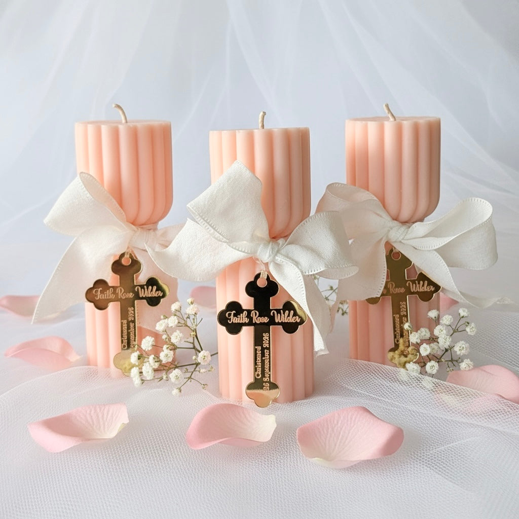 Three pastel pink ribbed candles tied with large white satin bows and decorated with gold cross charms, arranged on a white fabric surface with pink petals and small white flowers.