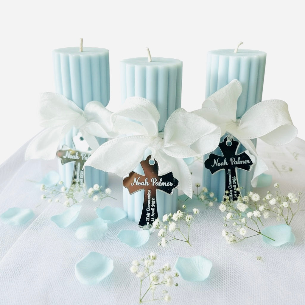 Three light blue pillar candles tied with large white satin bows, each decorated with a hanging cross tag and surrounded by white baby's breath flowers and pale blue petals on a white fabric surface.