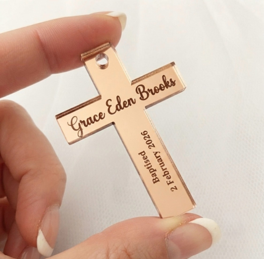 A close-up of a hand holding a personalized rose gold cross pendant engraved with the name 'Grace Eden Brooks' and baptism date '2 February 2026' against a soft white fabric background.